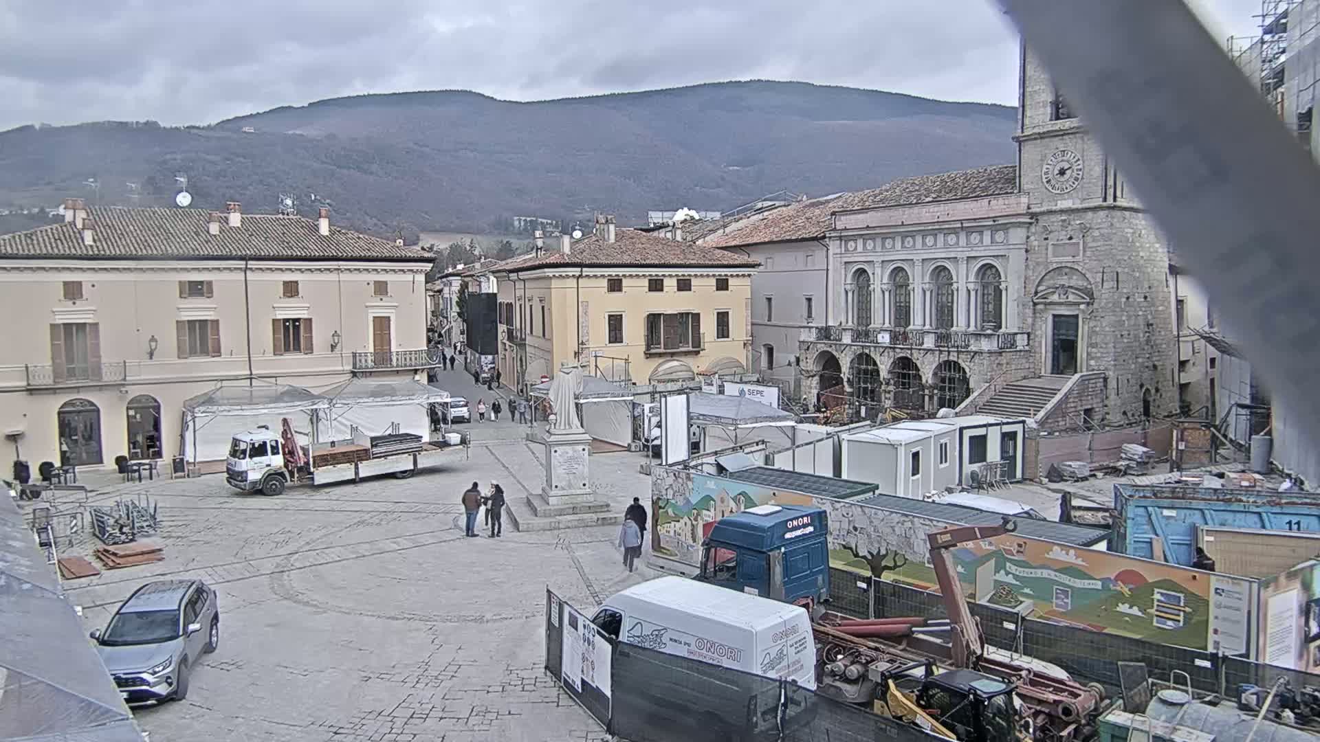 A town square under an overcast sky is undergoing construction, with trucks, temporary structures, and construction equipment visible.