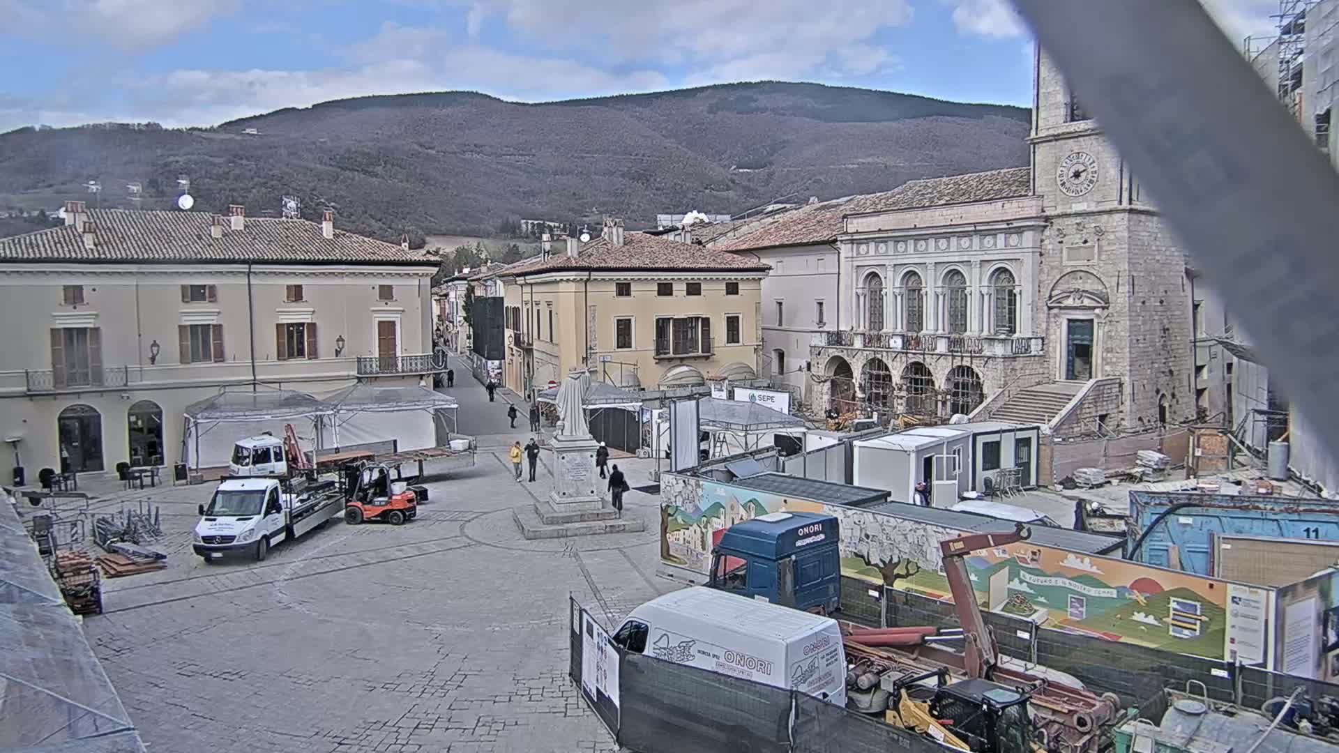 A town square under a partly cloudy sky is partially under construction, with several trucks and construction equipment present, and buildings surround the square, including one with a clock tower.