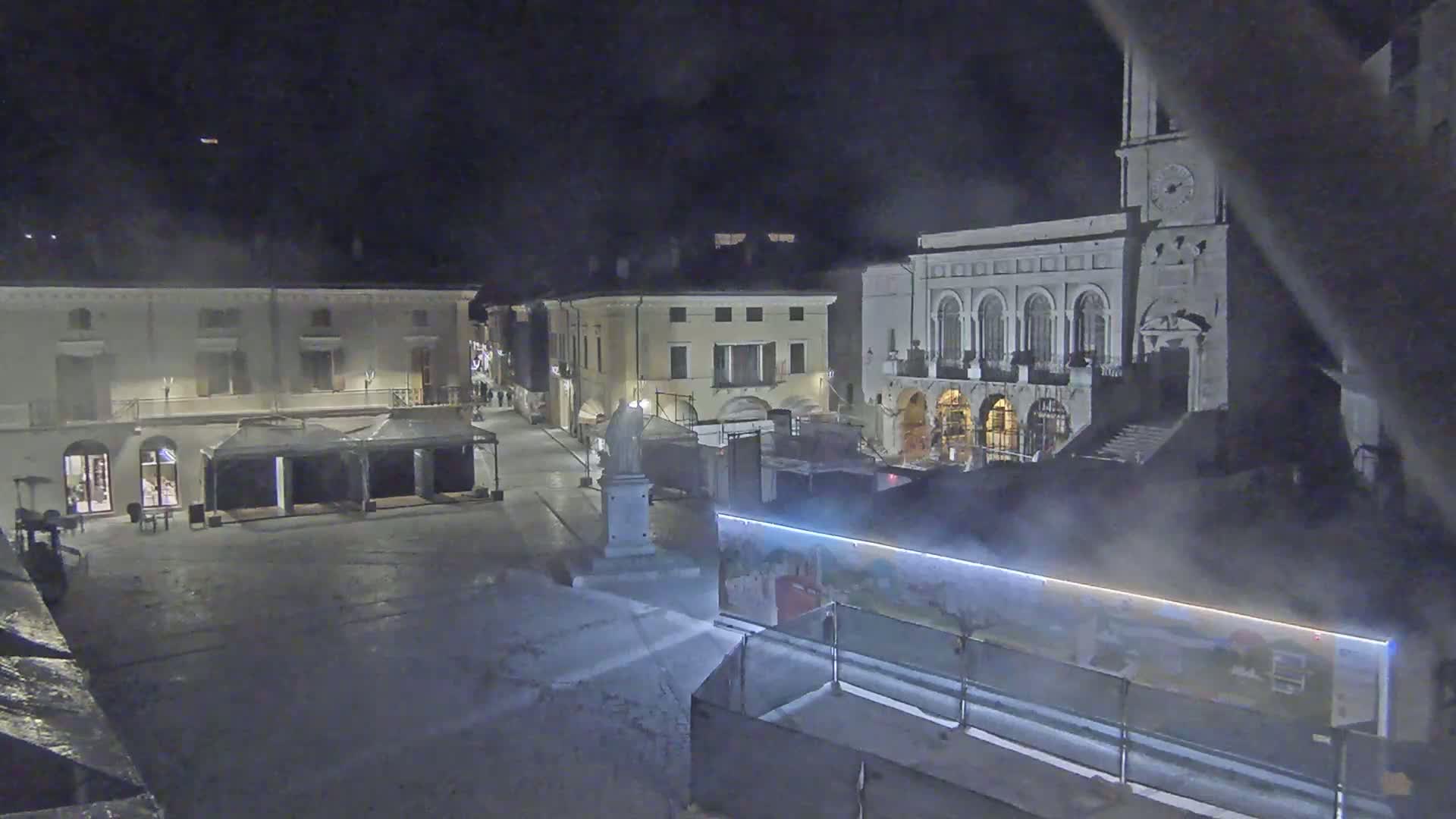 A nighttime view of a town square, with several buildings and a large construction barrier, showing some light mist or fog in the air.