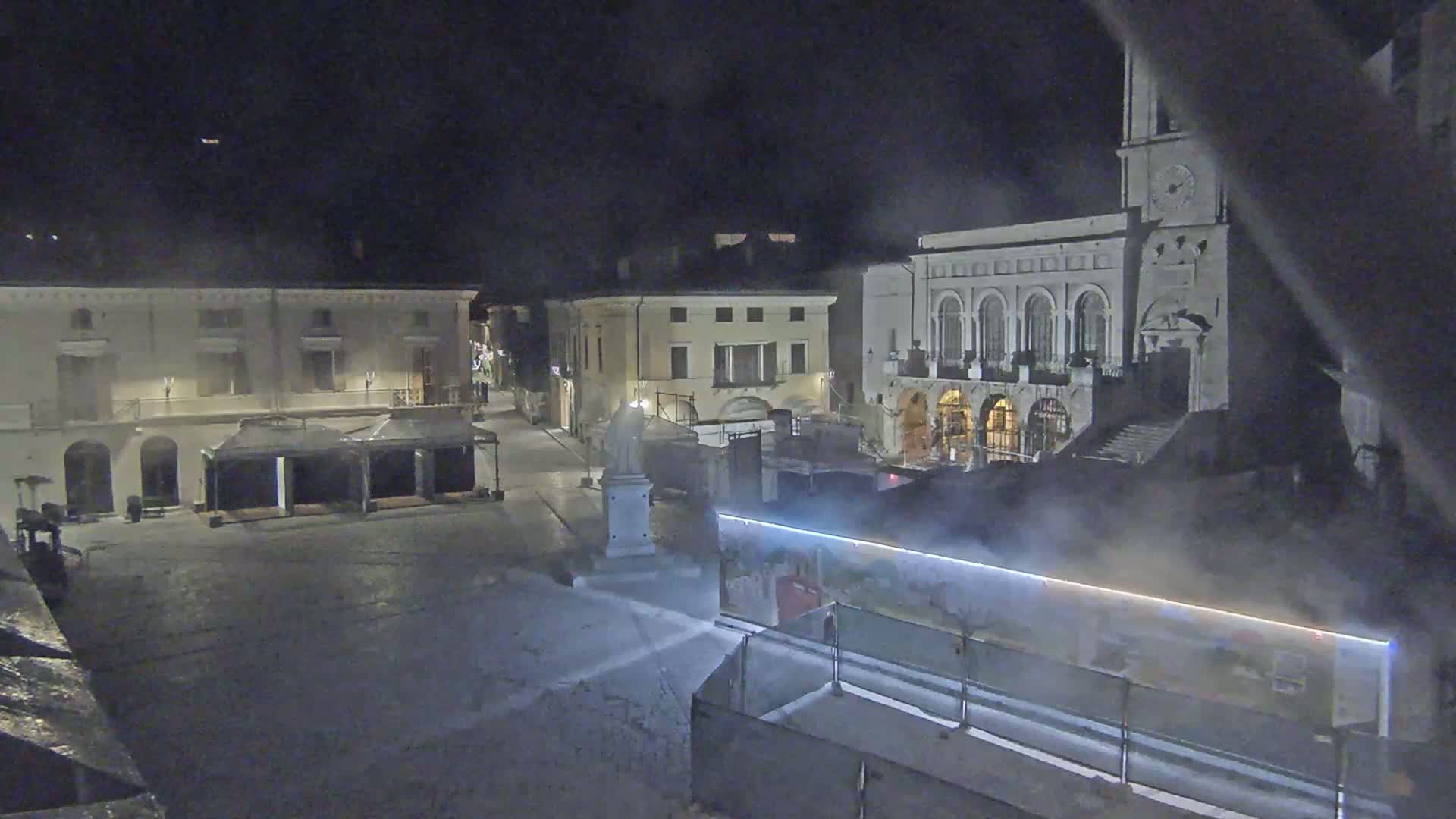 A nighttime view of a town square with several buildings, including one with a clock tower, partially obscured by mist or fog.