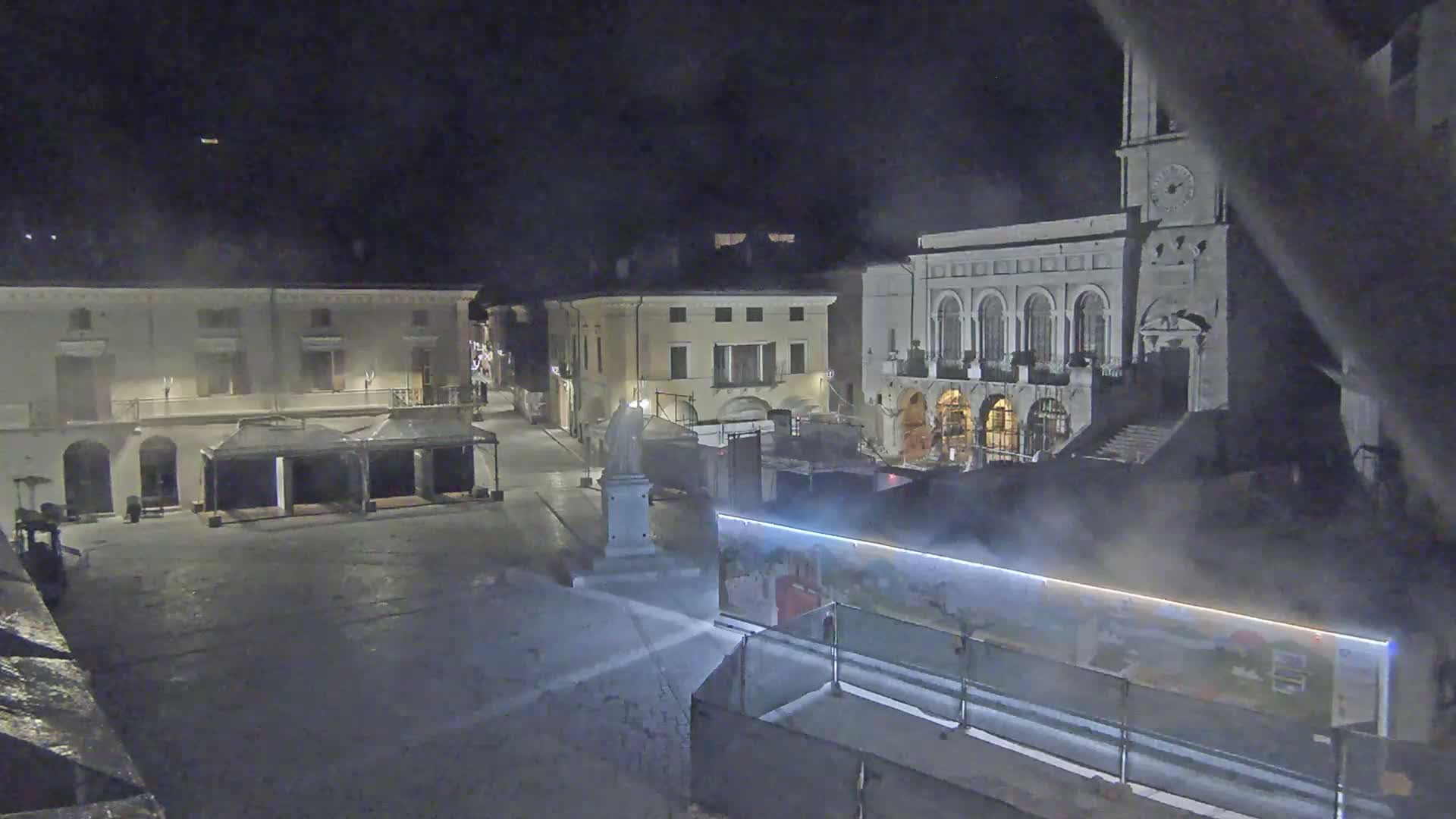 A nighttime view of a town square with several buildings, including a large building with arched windows and a clock tower, under hazy conditions.