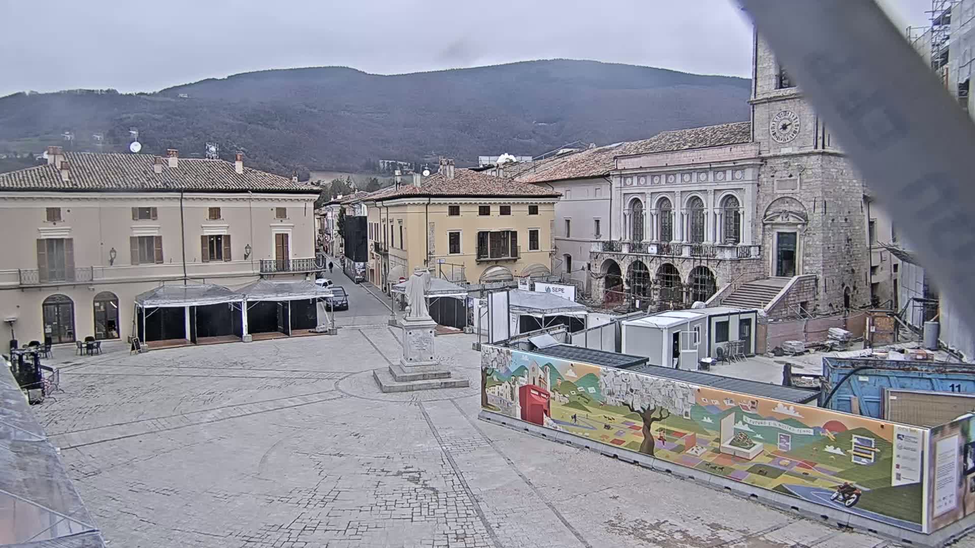 A town square with several buildings, some temporary structures, and construction materials is visible under an overcast sky, with mountains in the background.