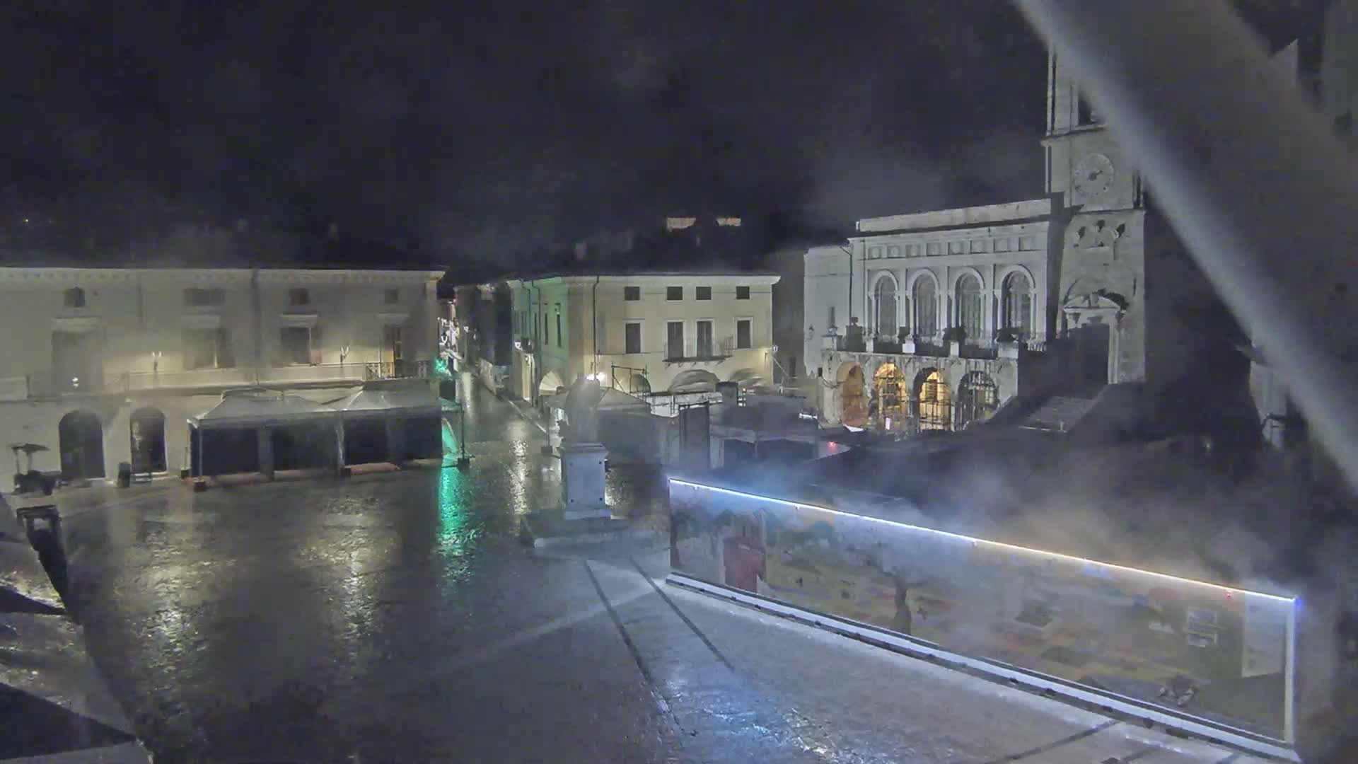 A rain-soaked town square at night is seen from above, with buildings surrounding it and steam rising from a structure in the foreground.