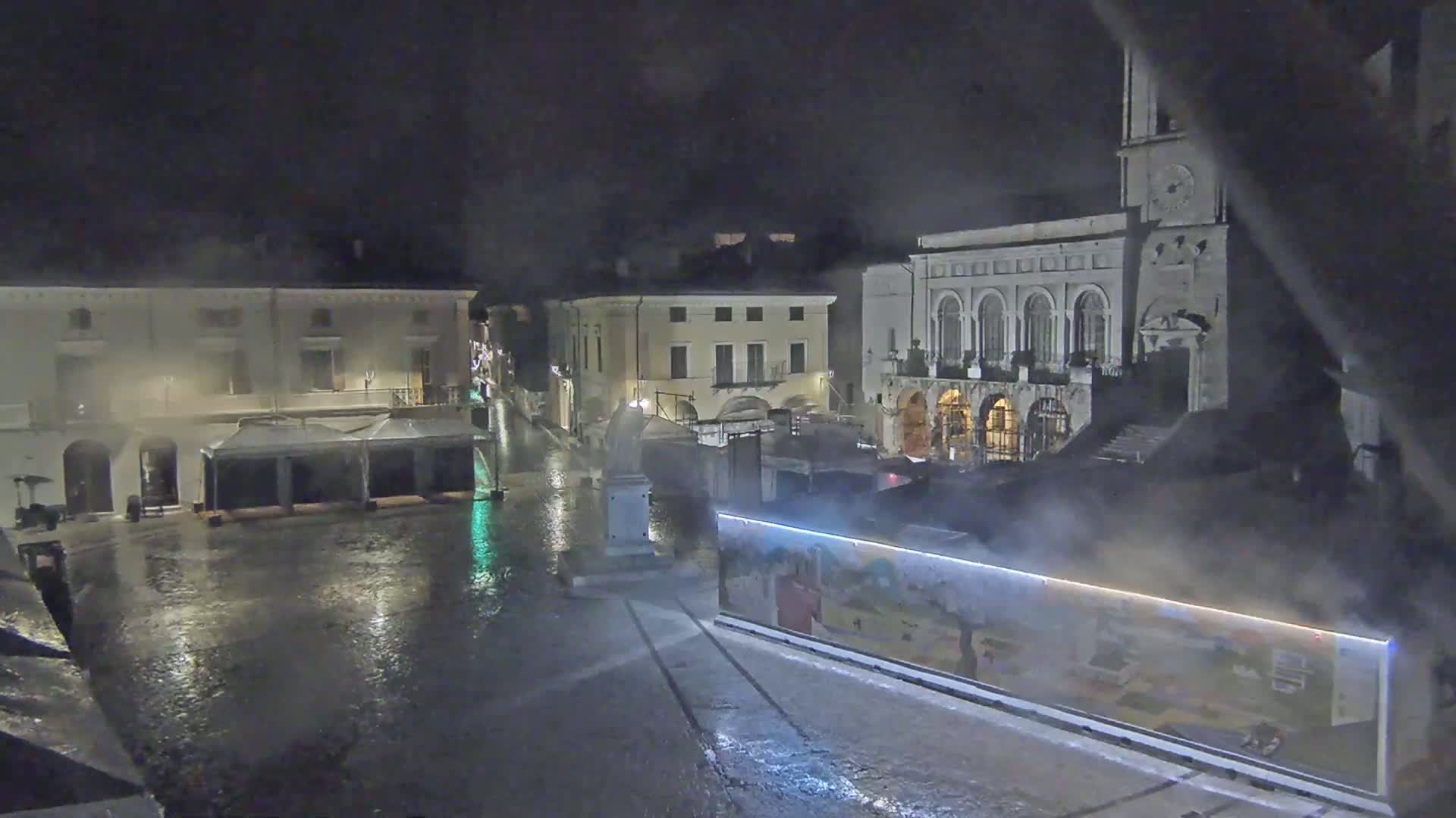 A rain-soaked town square at night, surrounded by buildings, one of which is a large structure with arched windows and a clock tower.