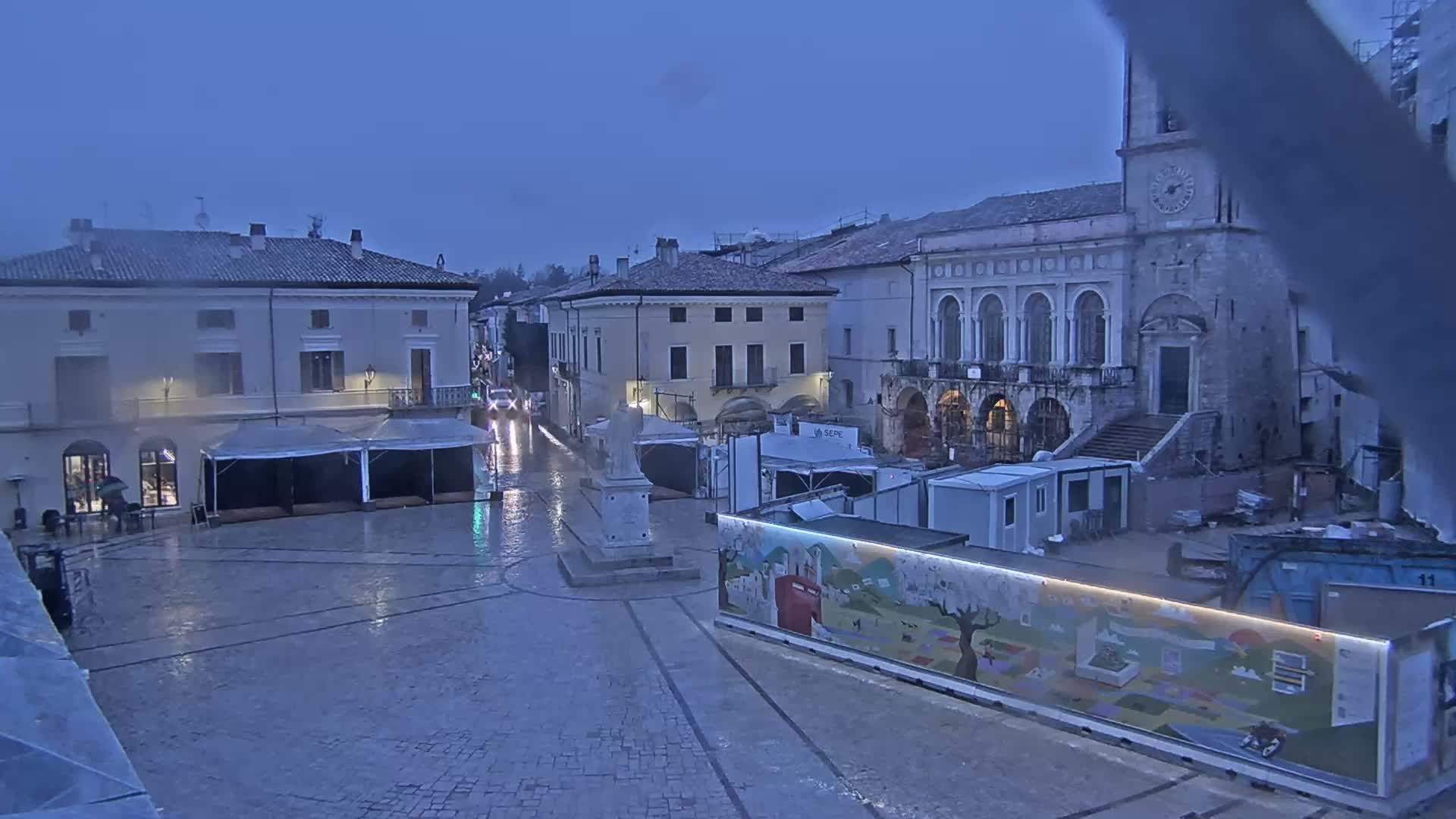 A rain-soaked town square, featuring several buildings, temporary structures, and a statue, is visible under a gloomy sky.