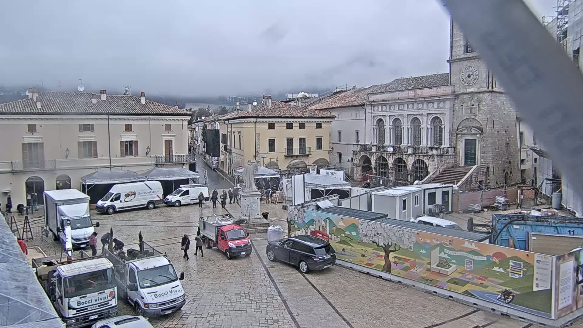 A town square with several trucks, temporary structures, and construction barriers is seen on an overcast day.