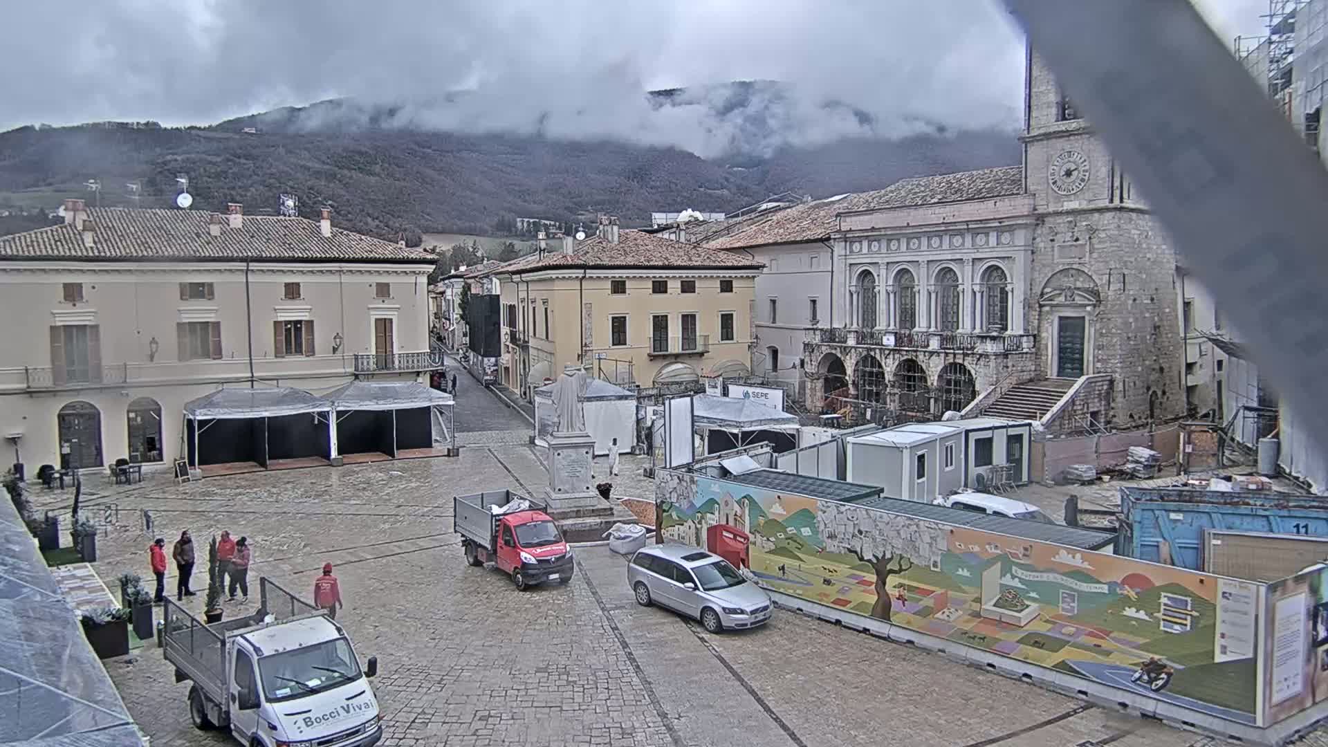 A town square under an overcast sky, partially obstructed by construction and temporary structures, is situated against a backdrop of misty mountains.