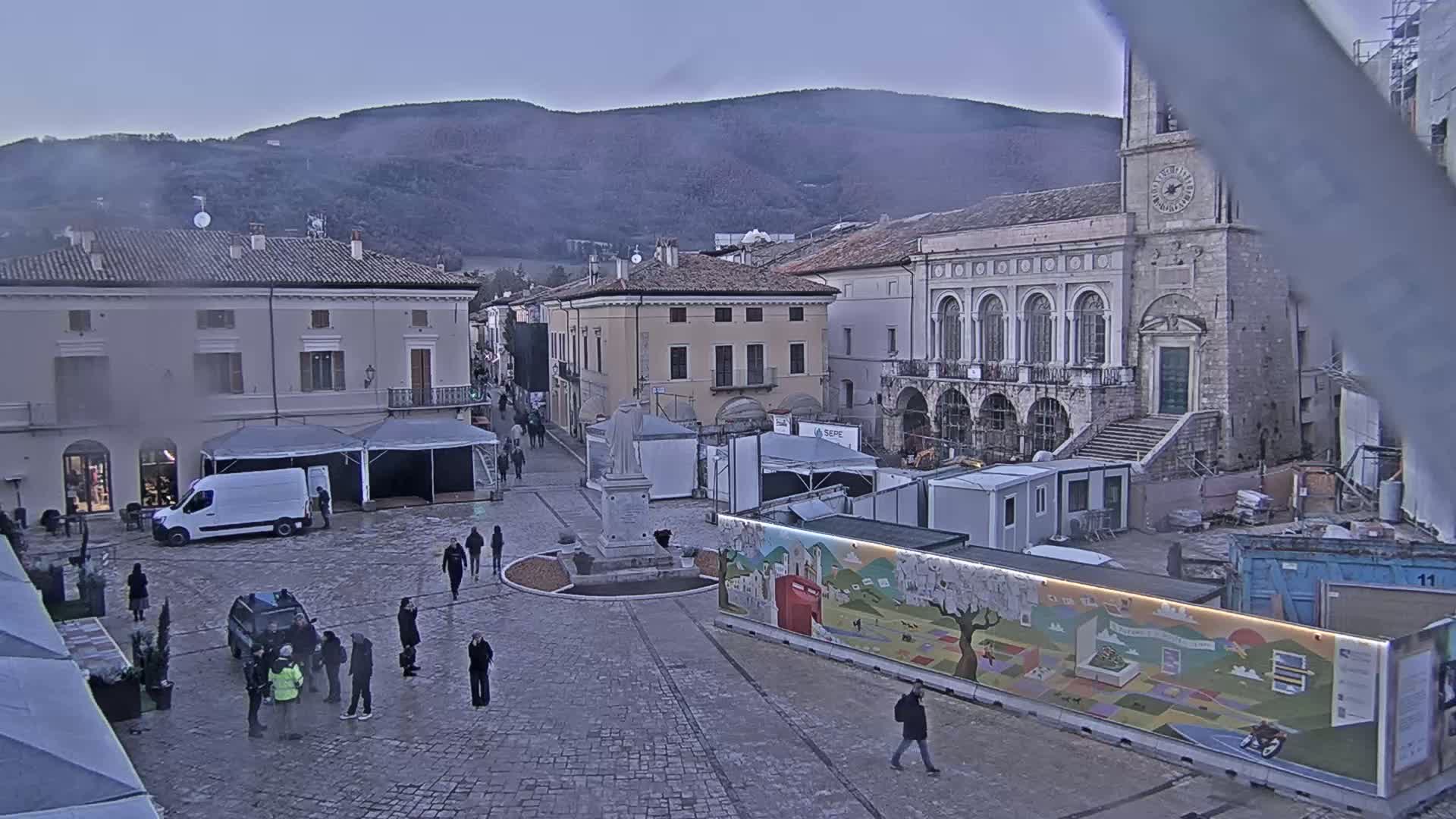 A town square with several temporary structures and a few people milling about under a dusky sky, with mountains visible in the background.