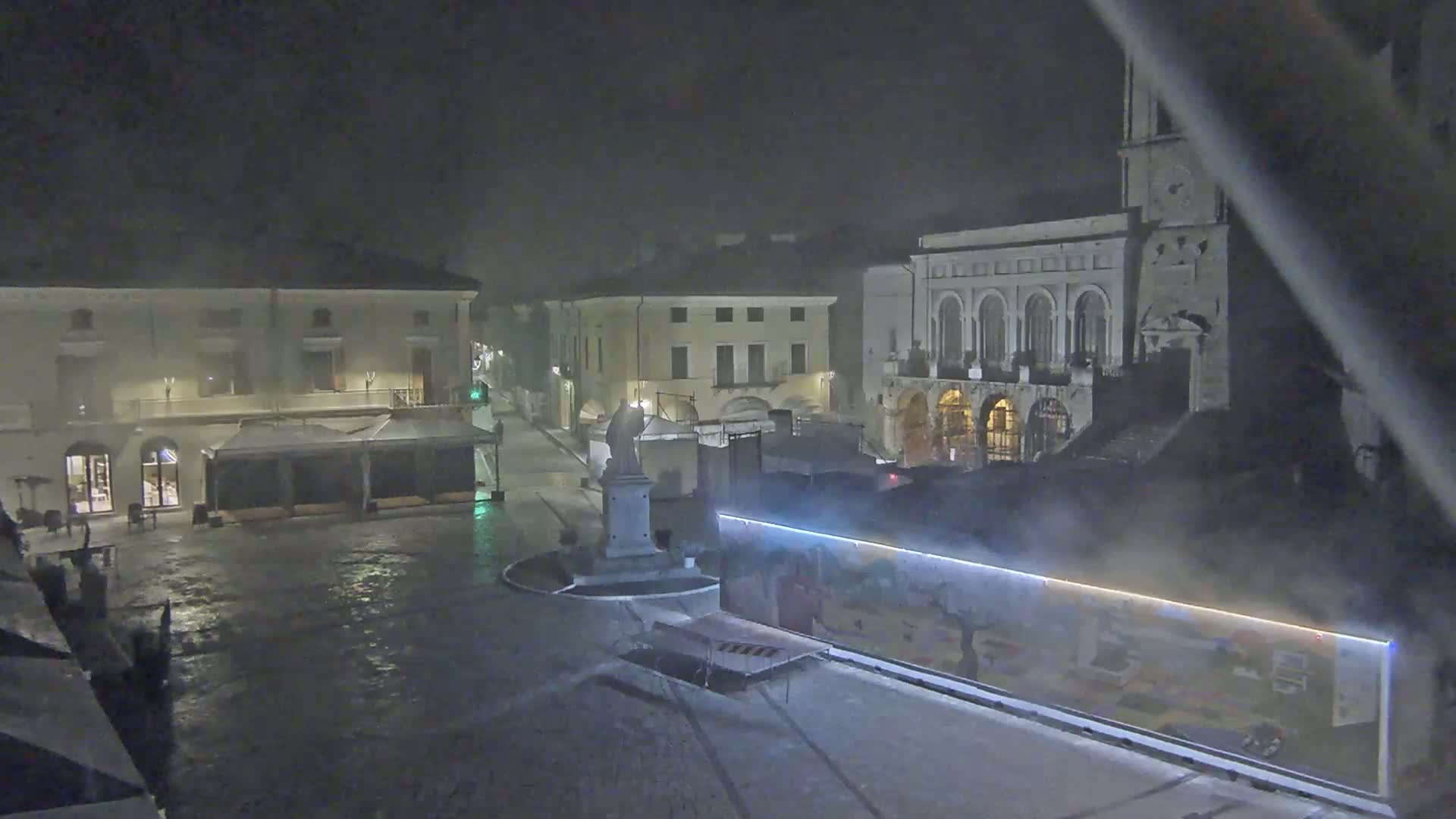 A wet, nighttime town square is seen with several buildings, including a large stone structure with arches and a statue on a pedestal in the center of the square.