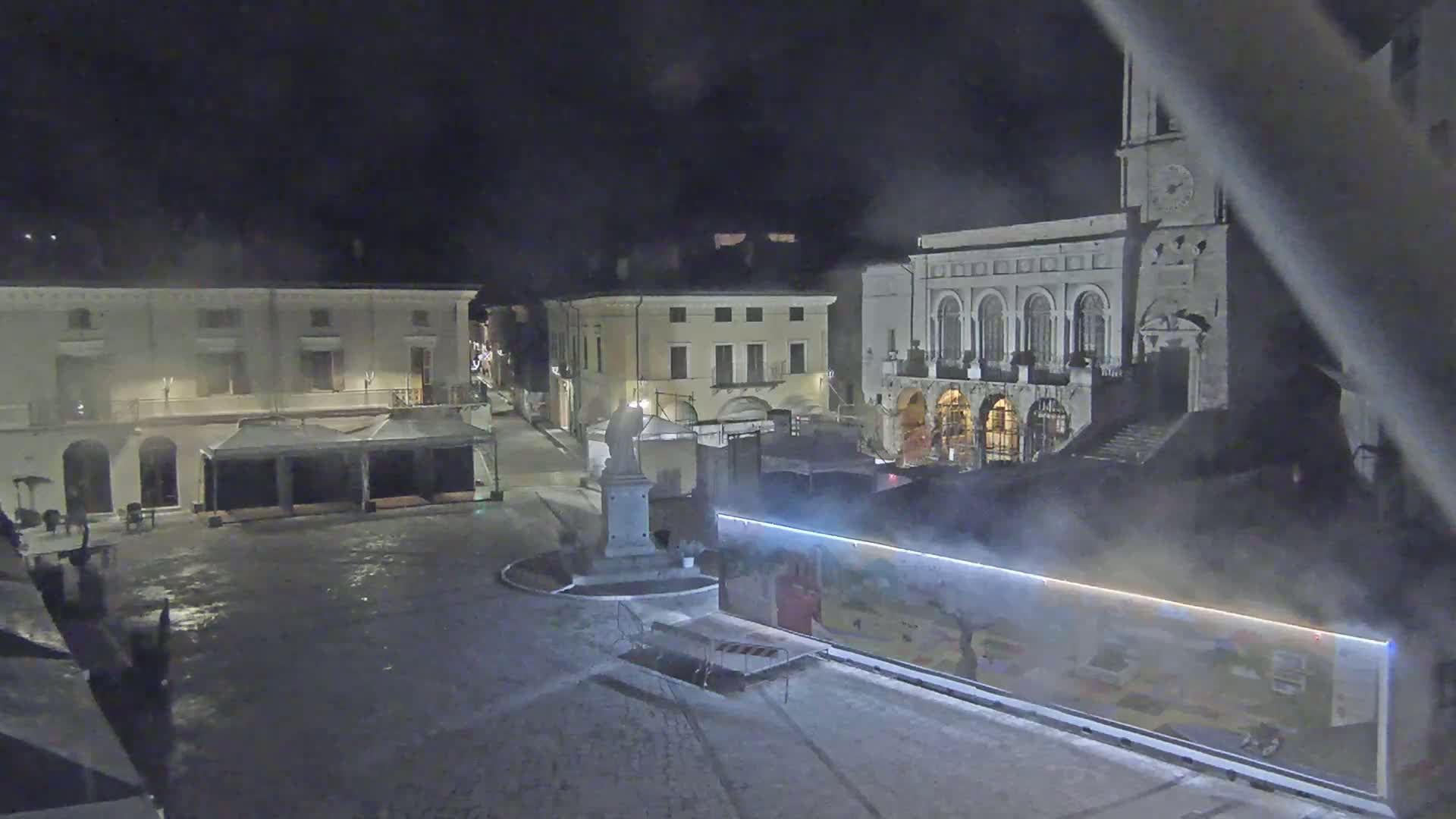 A nighttime view of a town square with several buildings, a statue, and temporary structures, under misty or foggy conditions.