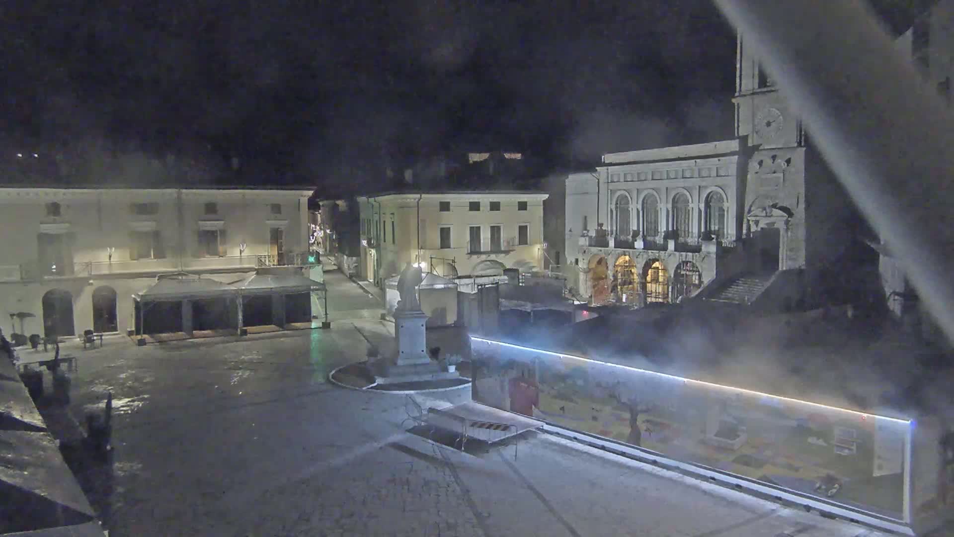 A nighttime view of a town square with several buildings, a statue, and temporary structures, under hazy or foggy conditions.