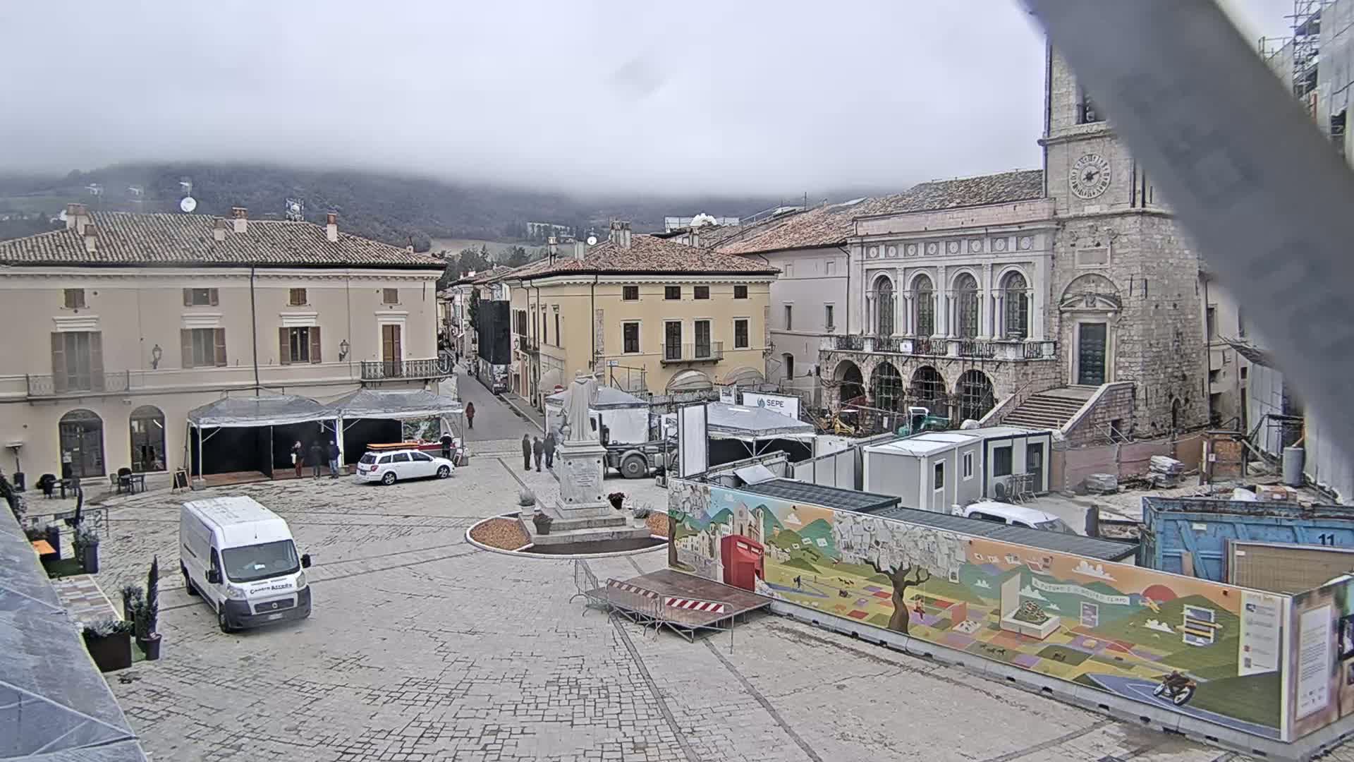 A town square in a European-style town, under an overcast sky, is partially blocked off by construction and temporary structures, with a few people and vehicles present.