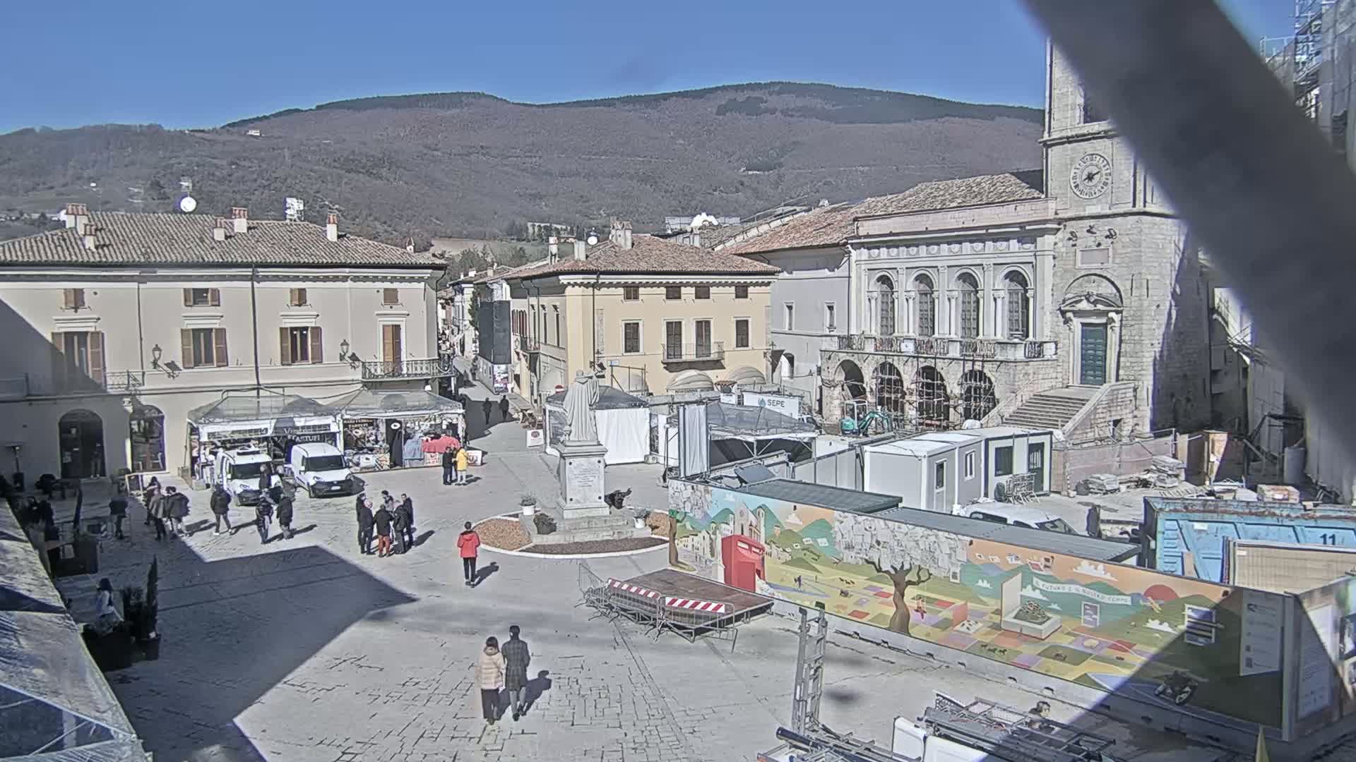 A town square with several buildings, temporary stalls, a statue, and construction materials is visible on a sunny day with mountains in the background.