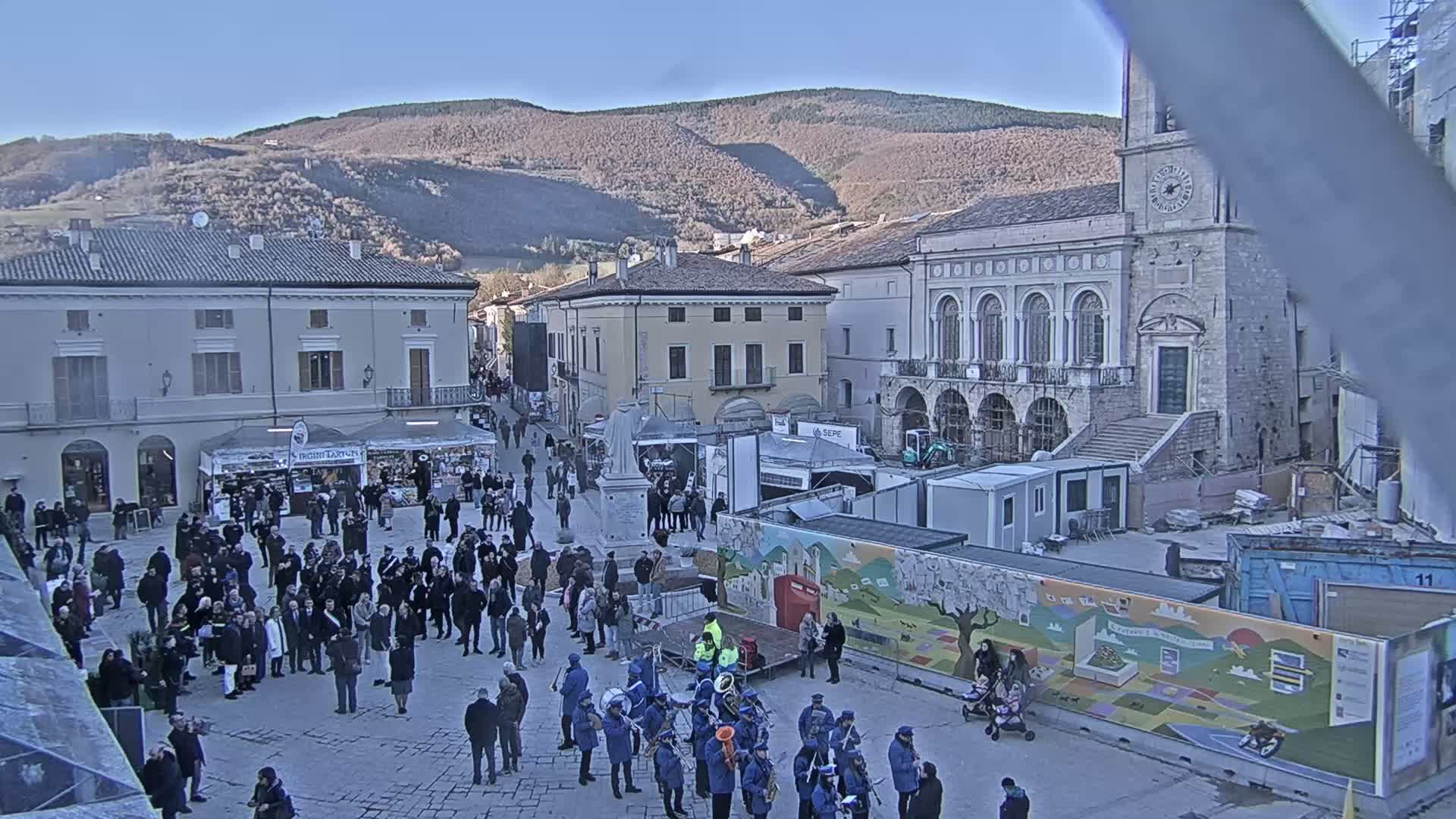 A large crowd gathers in a town square on a sunny day, with temporary market stalls and a brass band, in front of historic buildings and mountains in the background.