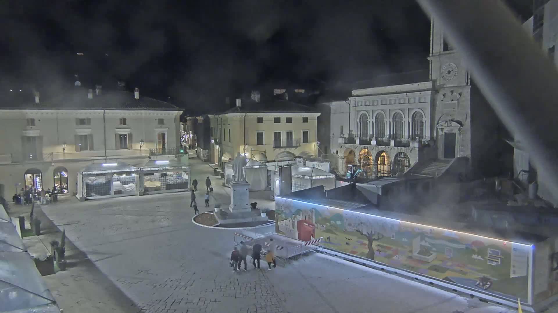 A nighttime view of a town square with several temporary stalls and a statue, under a hazy sky.