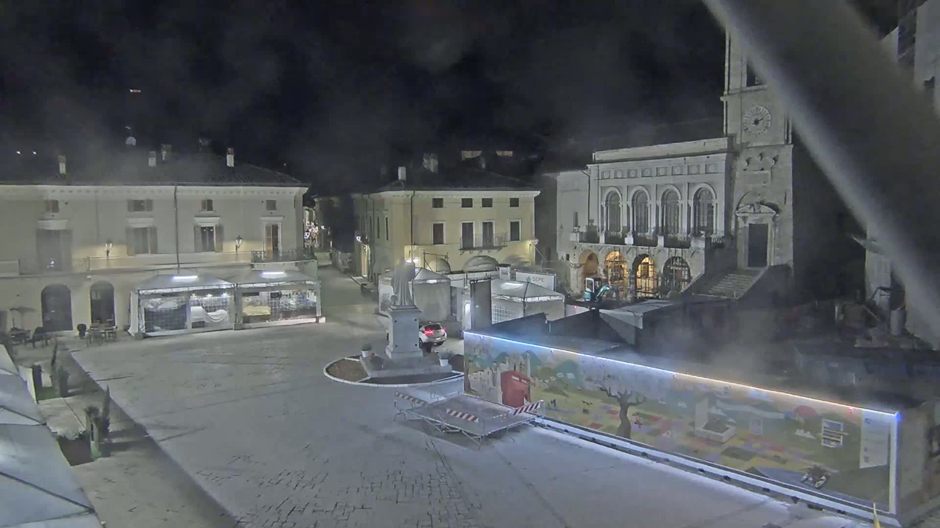 A nighttime view of a town square, with several buildings and temporary structures, under hazy or foggy conditions.