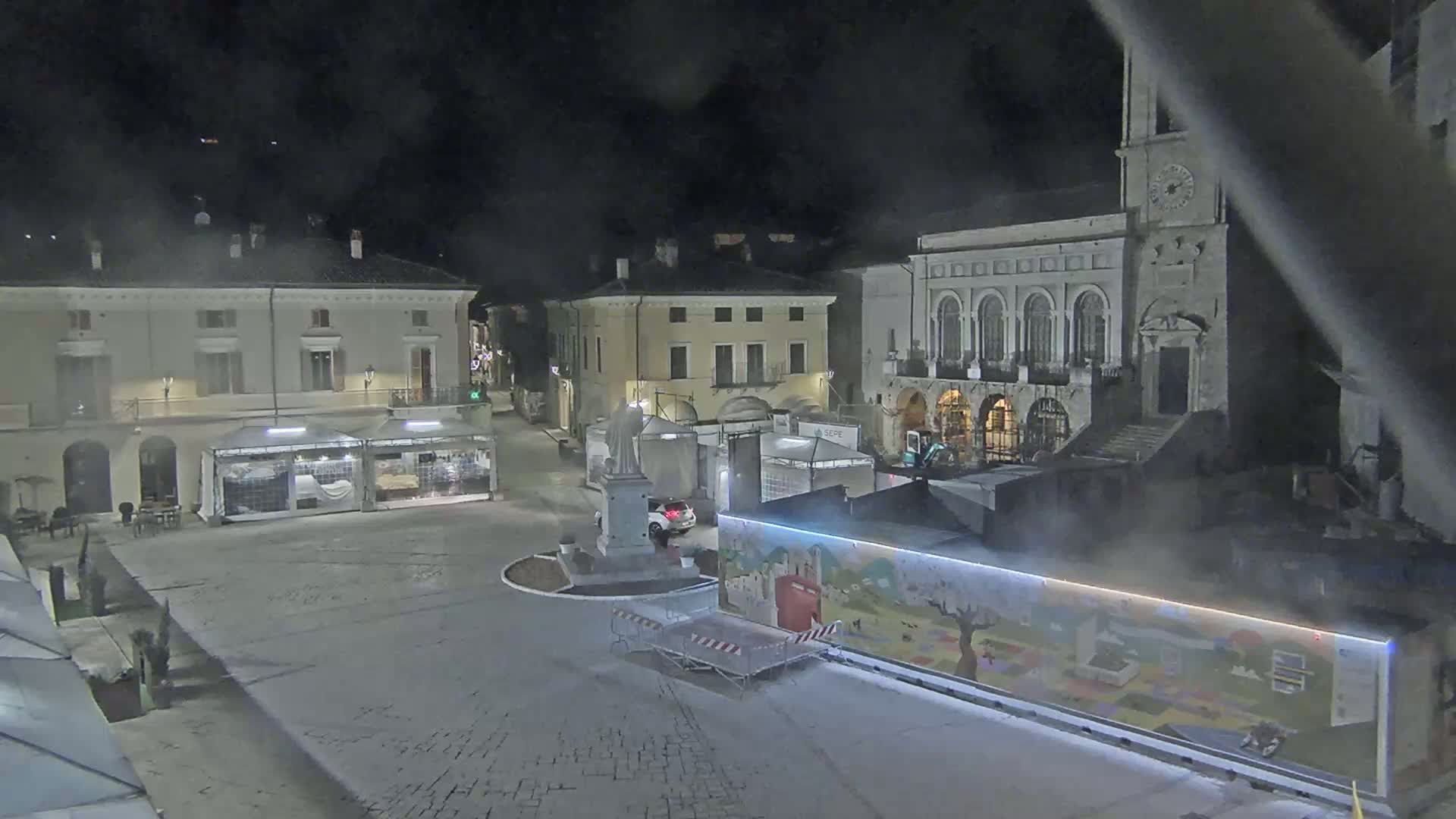 A nighttime view of a town square with several buildings, temporary market stalls, and a large mural-covered structure, under hazy or foggy conditions.