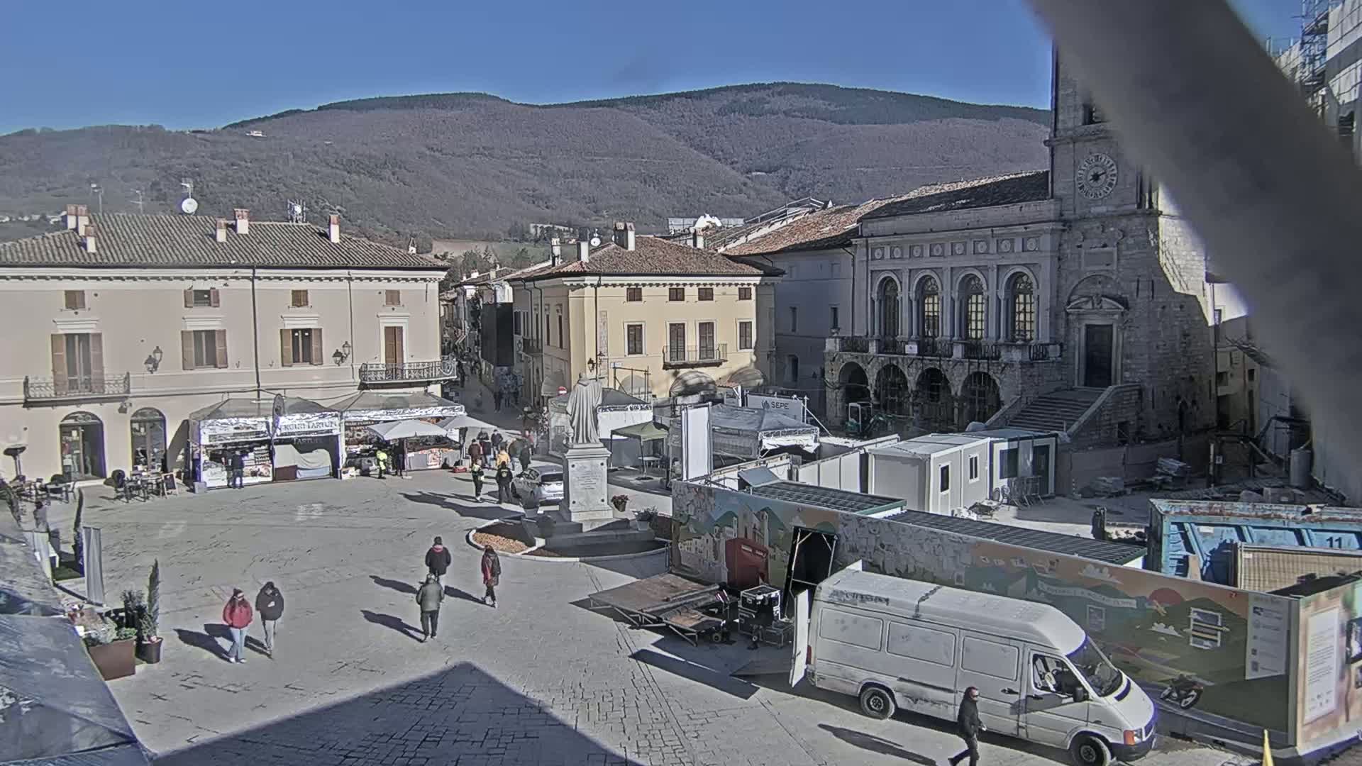 A town square with several buildings, market stalls, a statue, and temporary structures, under sunny skies and in front of a mountain range.