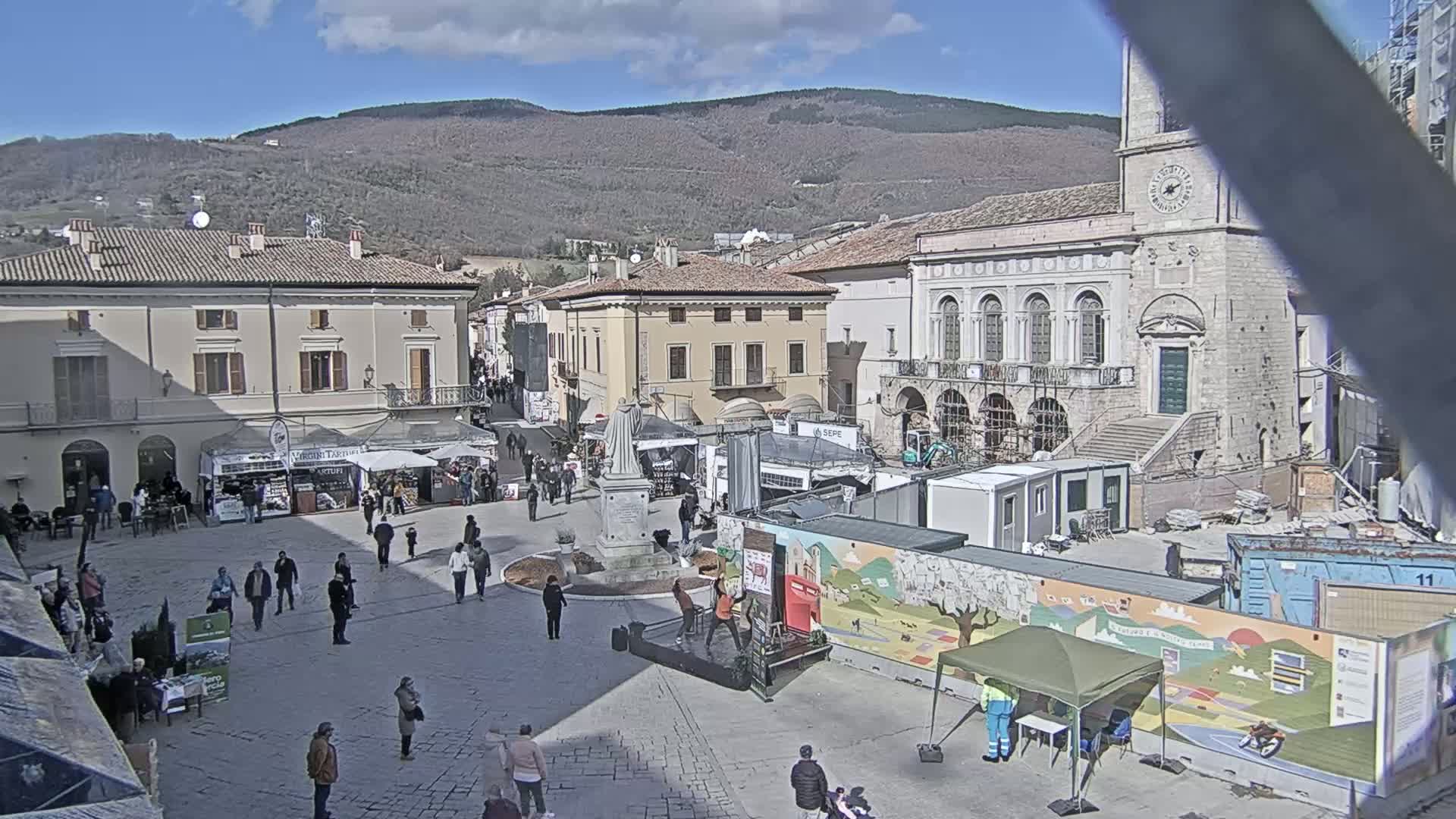 A town square filled with people and market stalls under a sunny sky, with mountains in the background and construction work underway near a large stone building.