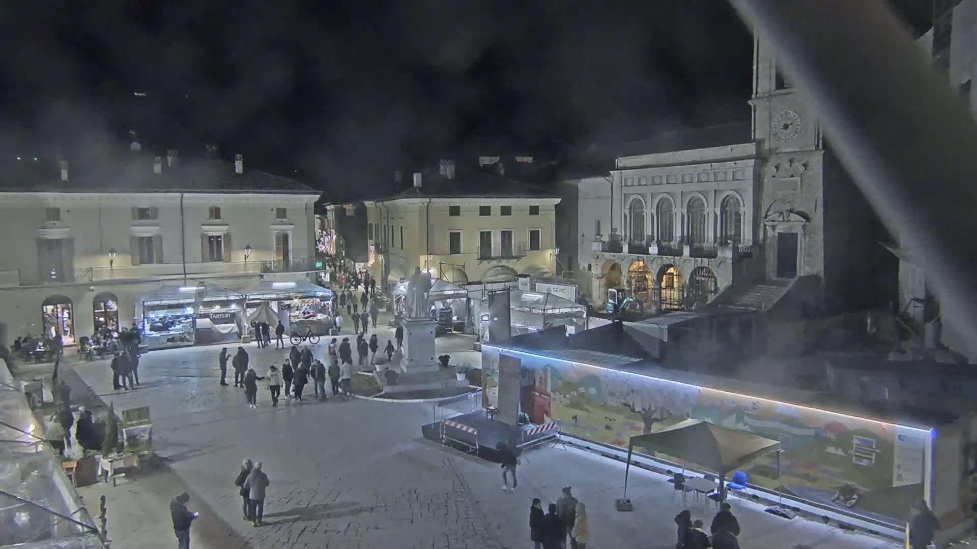 A nighttime town square with several market stalls and people milling about, under a hazy, dark sky.
