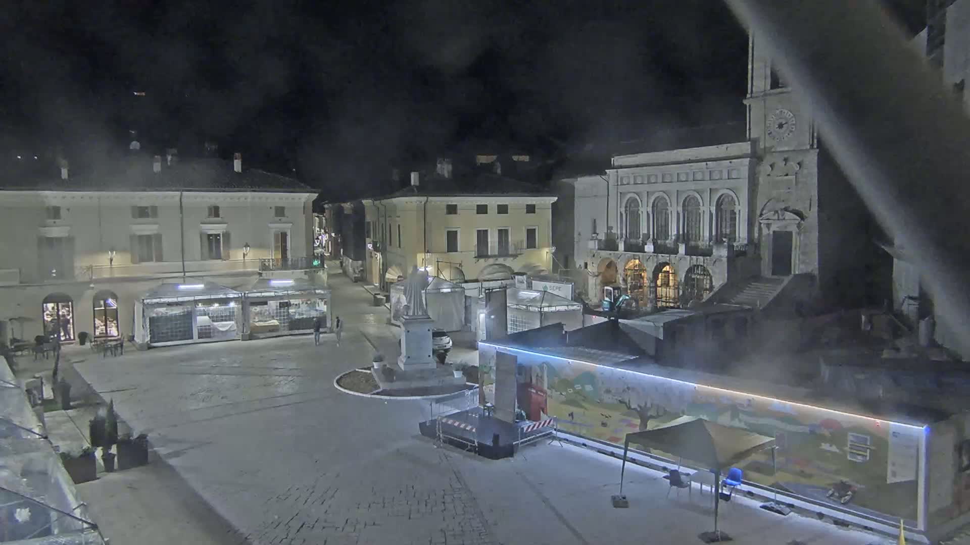 A nighttime view of a town square with several buildings, temporary stalls, and a stage setup, under hazy or foggy conditions.