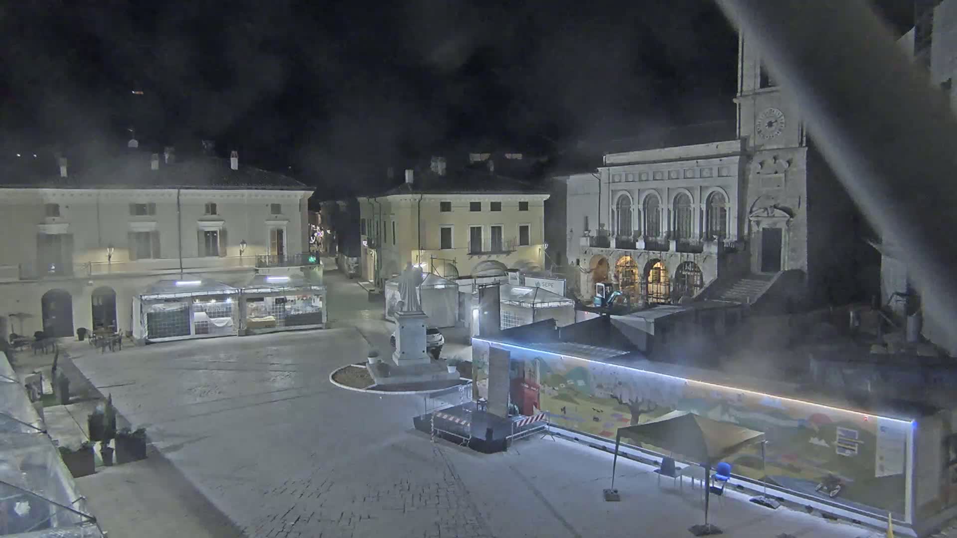 A nighttime view of a town square with several buildings, temporary market stalls, and a stage, under hazy or foggy conditions.