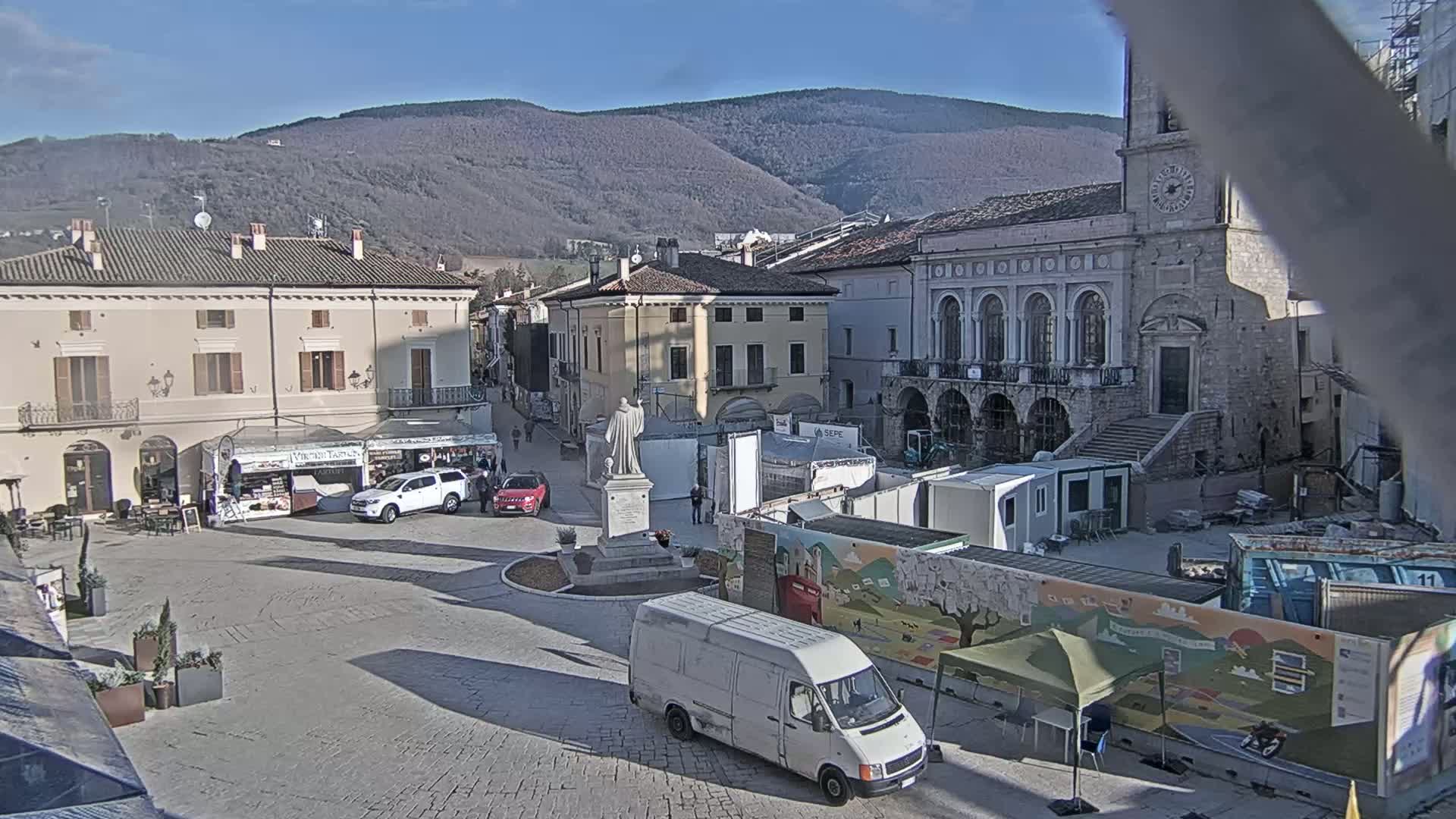 A town square with several buildings, a statue, some vehicles, and temporary market stalls under a clear, sunny sky and mountains in the background.
