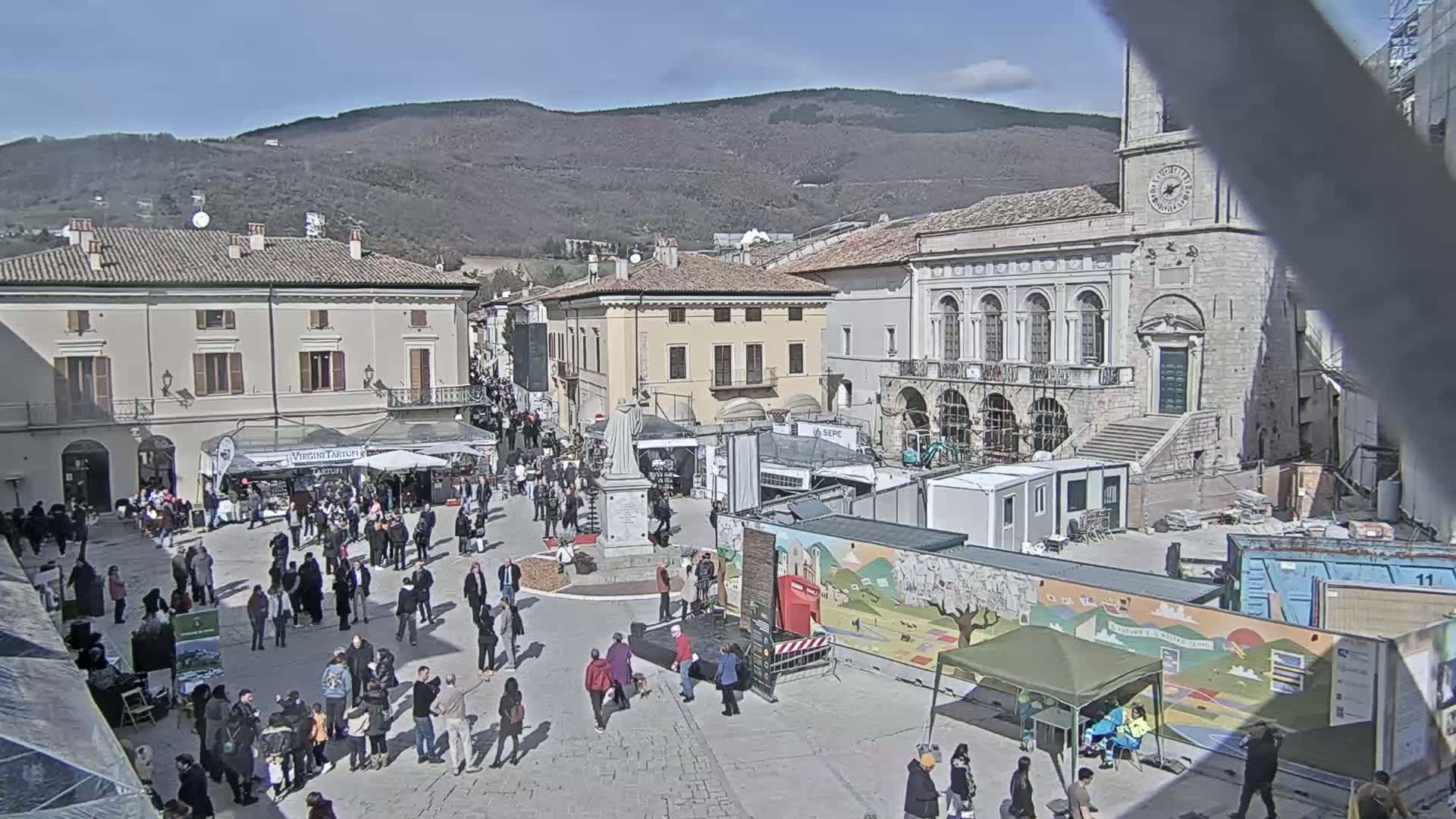 A sunny day in a town square, with many people gathered around market stalls and temporary structures near historic buildings under construction.