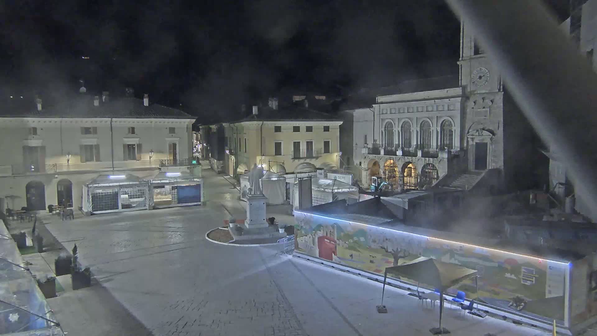 A nighttime view of a town square with several buildings, temporary stalls, and a statue, under a hazy or foggy sky.