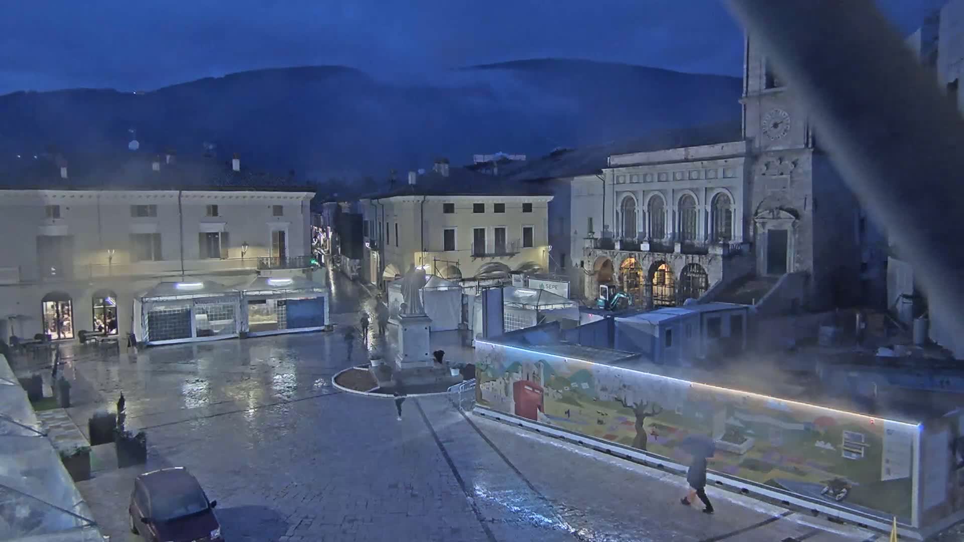 A rain-slicked town square at night, surrounded by buildings, with temporary structures and a few pedestrians visible under a dark sky.