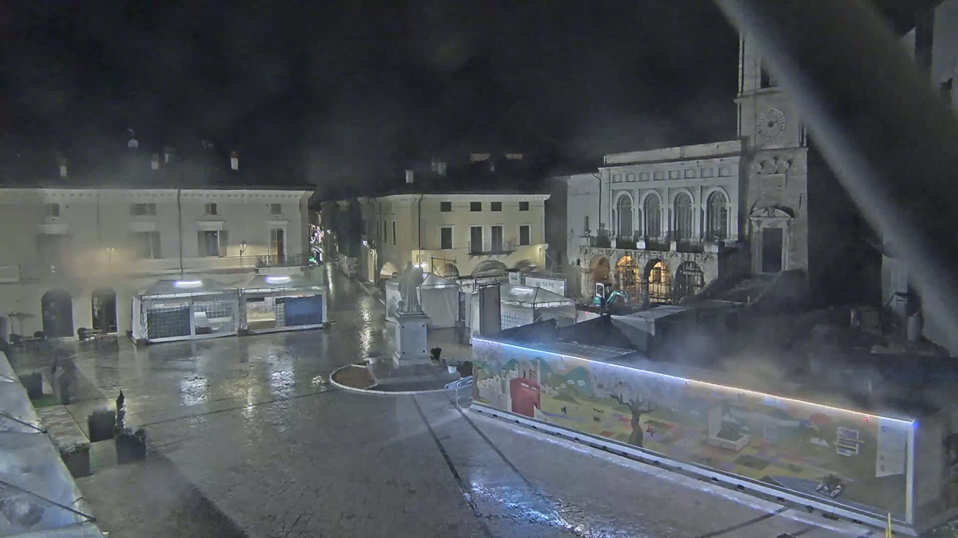 A rain-slicked town square at night features several temporary market stalls surrounding a statue and older buildings, one of which is a church.