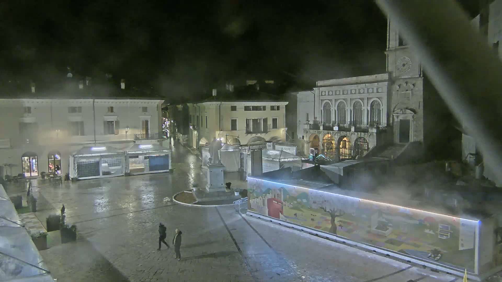 A wet, nighttime town square is sparsely populated with a few pedestrians, temporary market stalls, and buildings, one of which is a large stone structure with arched windows.