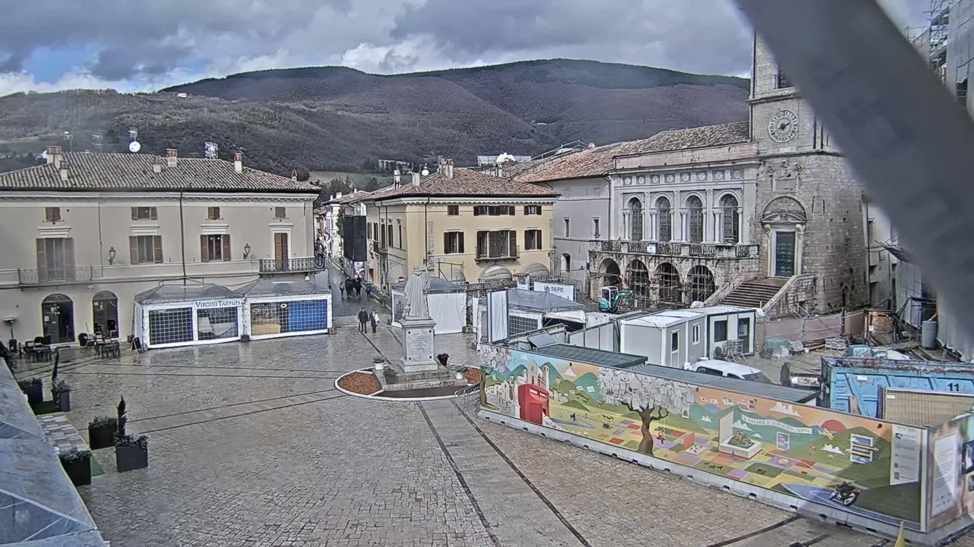 A town square with several buildings, a statue, temporary structures, and construction equipment is seen under an overcast sky, with mountains visible in the background.