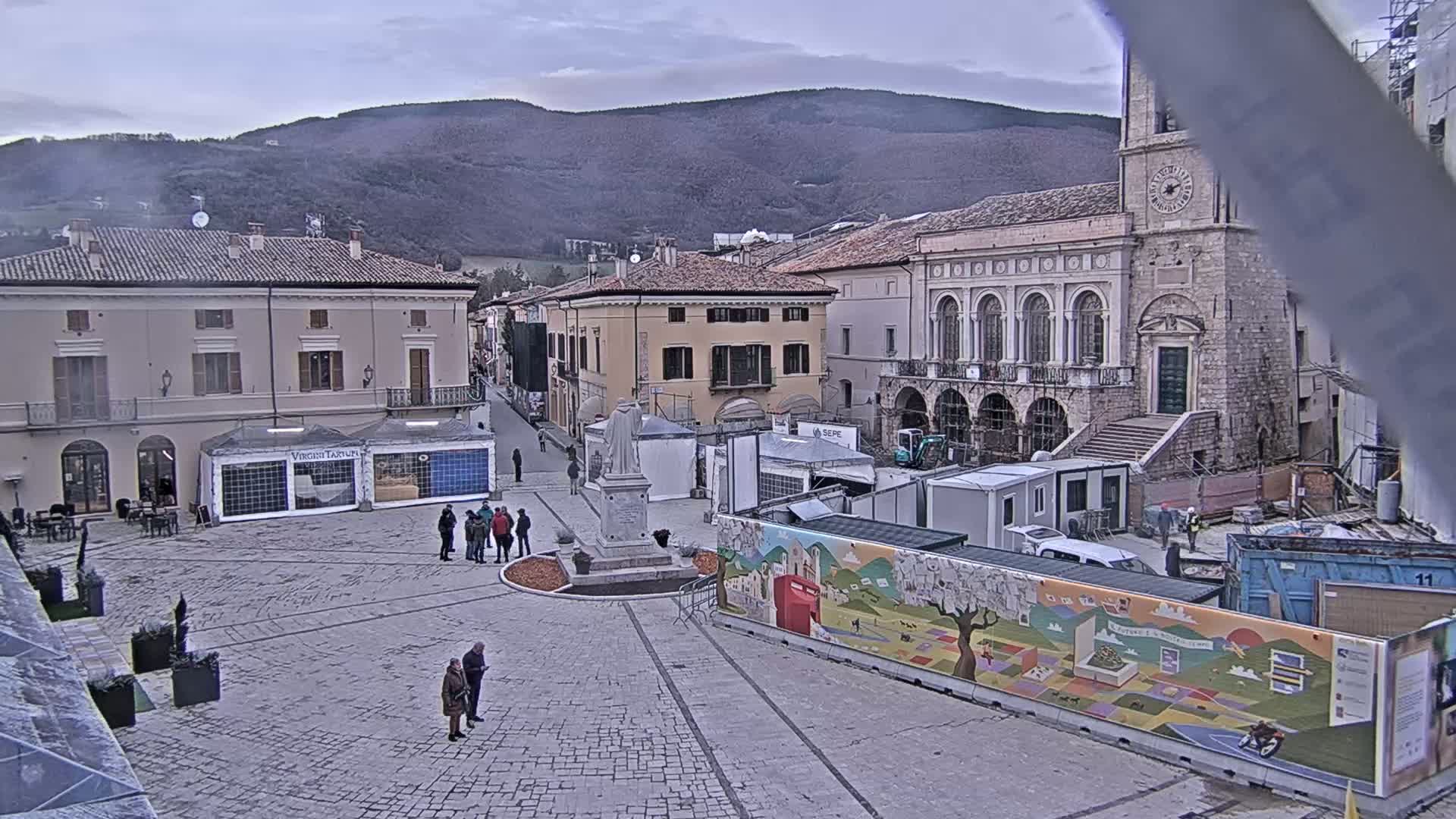 A town square, under an overcast sky, features several buildings, a statue, temporary structures, and a few people.