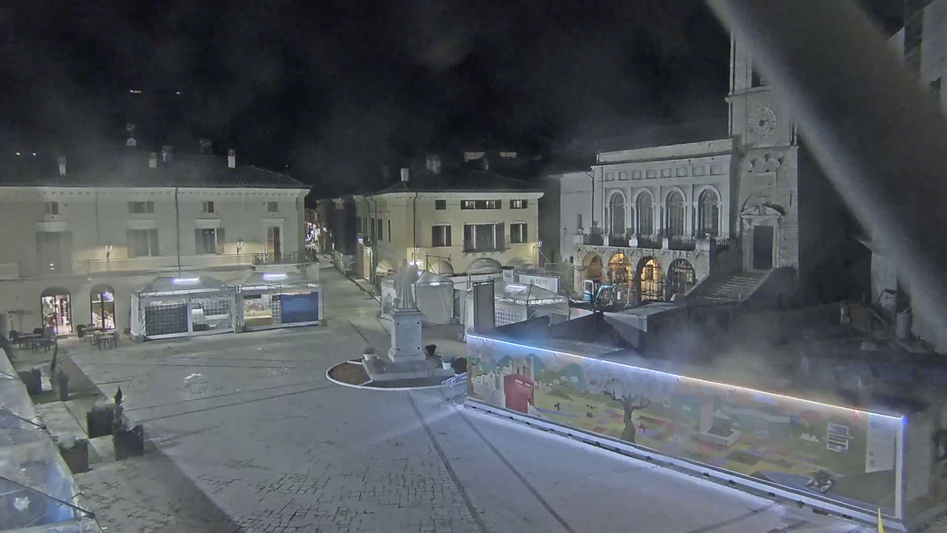 A nighttime view of a town square with several buildings, temporary stalls, and a statue, under hazy or foggy conditions.