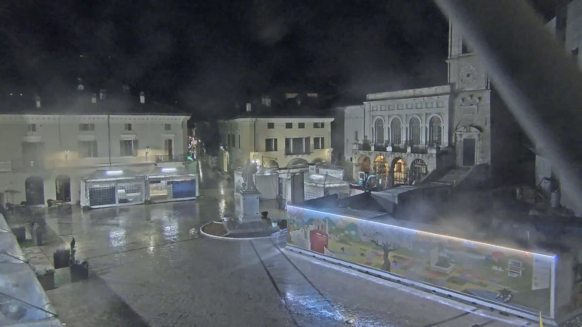 A rain-soaked town square at night, featuring several buildings, temporary market stalls, and a statue in the center.
