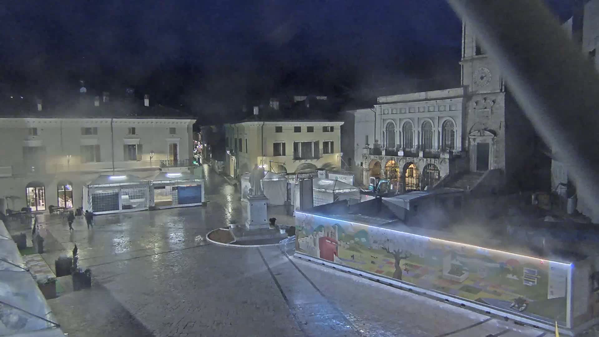 A wet, nighttime town square is seen with several temporary structures and buildings surrounding it, one of which has a clock tower.