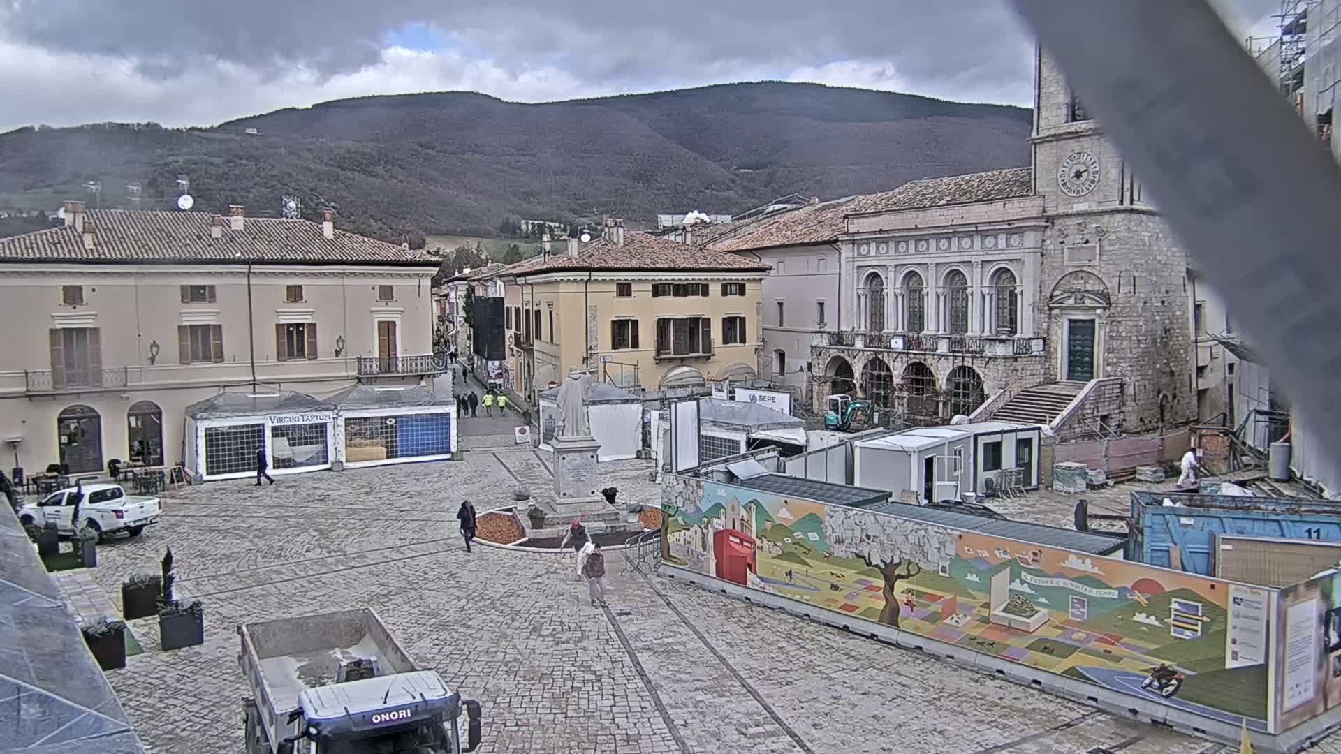 A town square under an overcast sky is being prepared for an event, with temporary structures, construction equipment, and a painted wall visible.