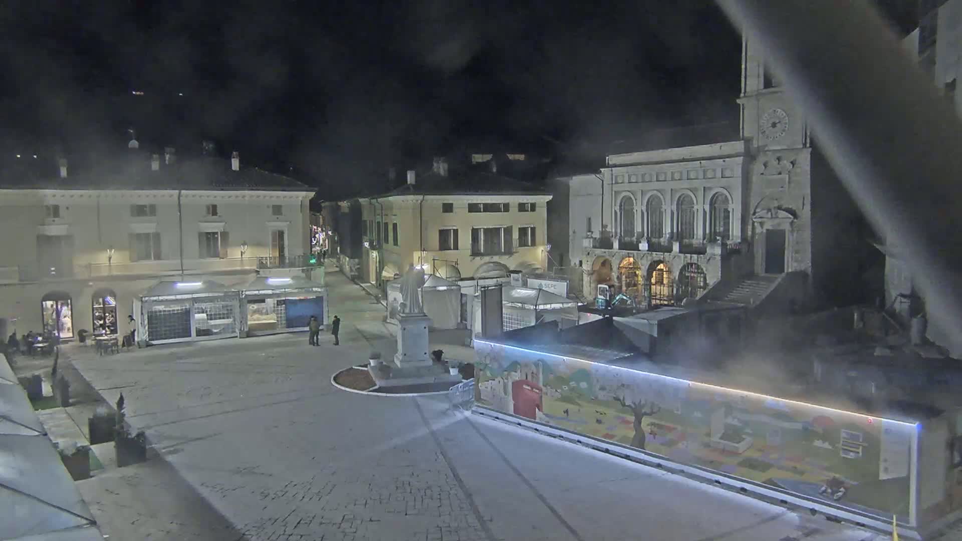 A nighttime view of a town square with several buildings, temporary market stalls, and a statue, under hazy or foggy conditions.