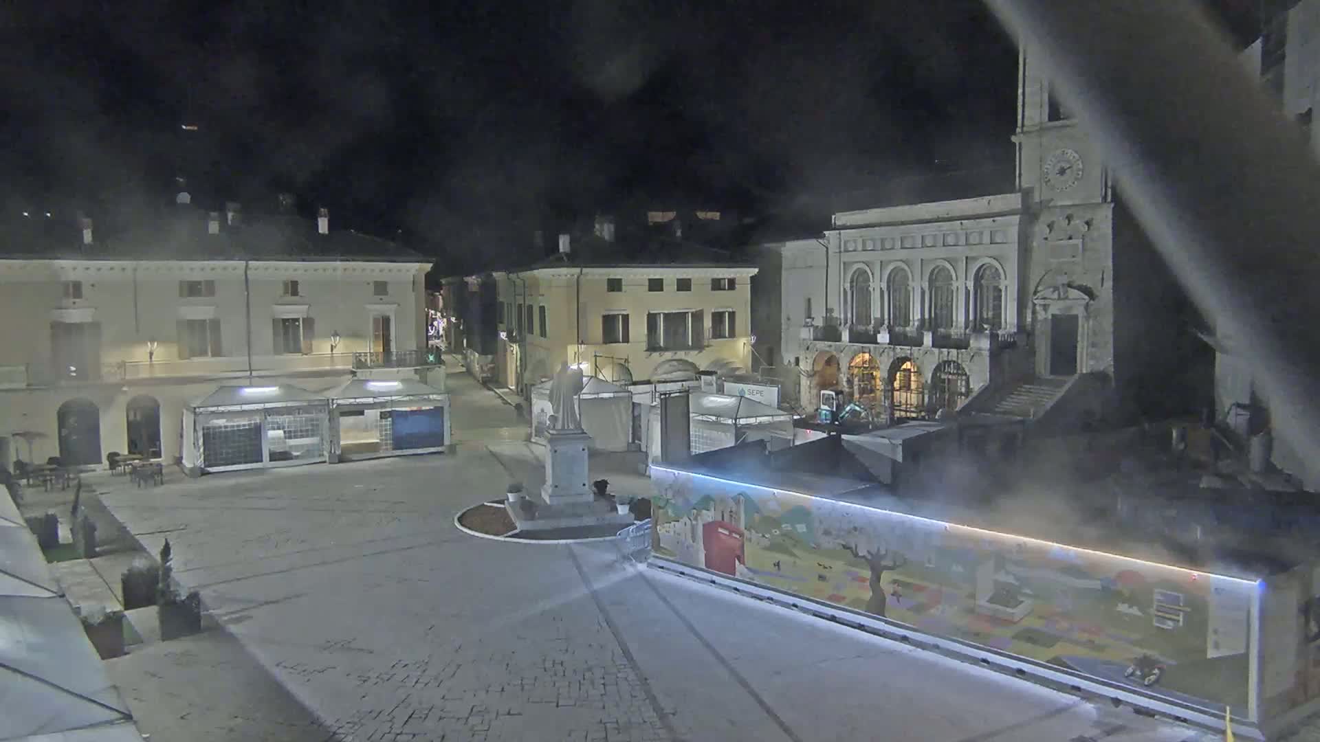 A nighttime view of a town square with several buildings, temporary market stalls, and a statue, with a light fog or mist present.