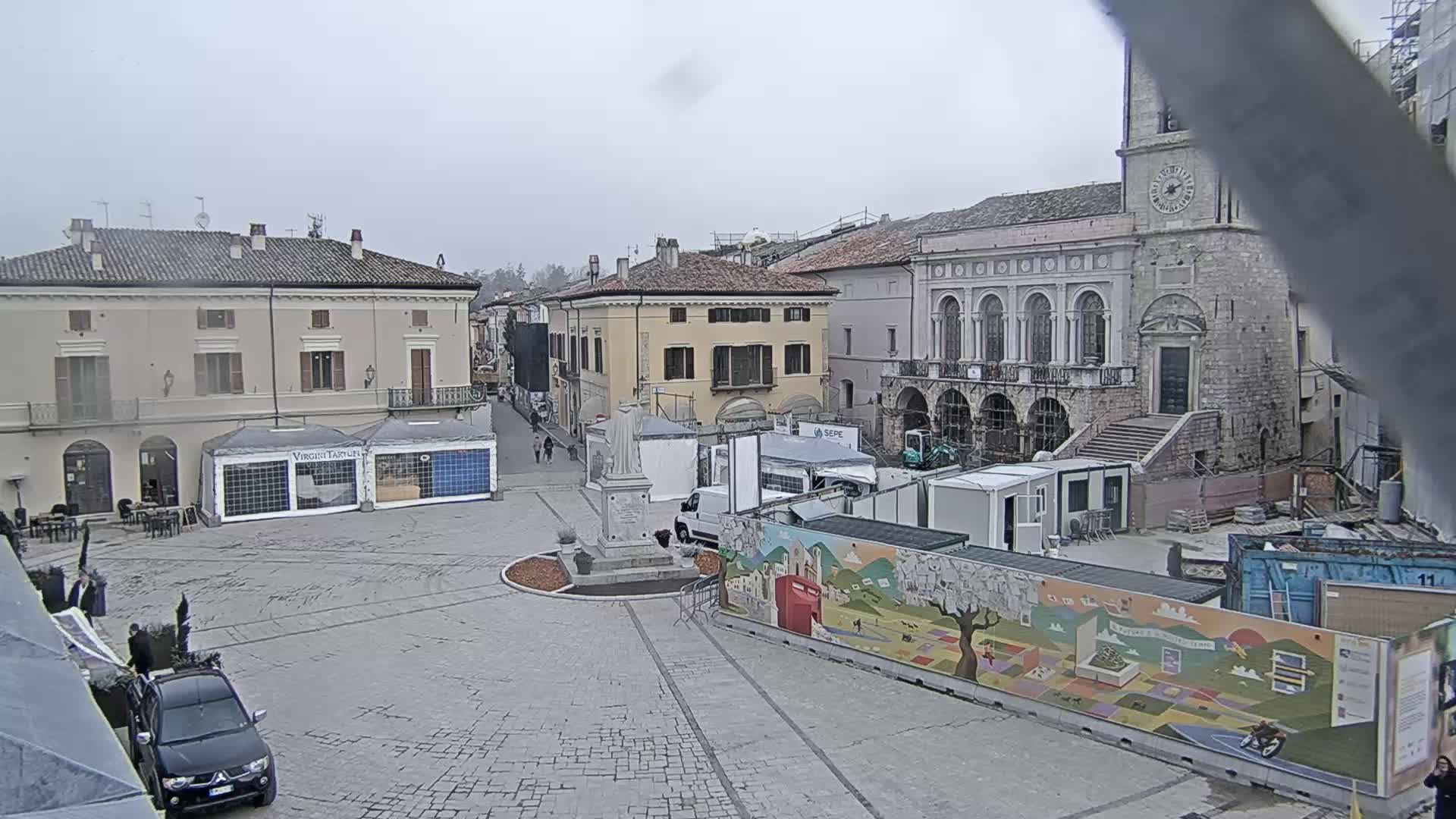 A town square with several buildings, temporary structures, and a statue, under an overcast sky.