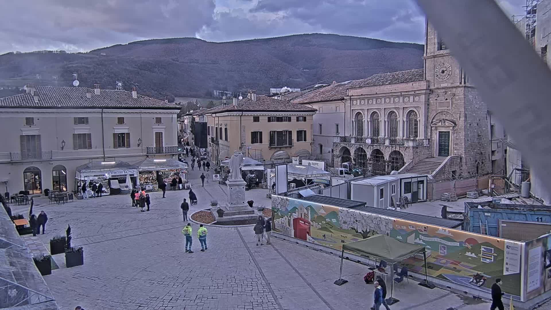 A town square with several temporary stalls and a statue, surrounded by buildings under an overcast sky, with mountains in the background.