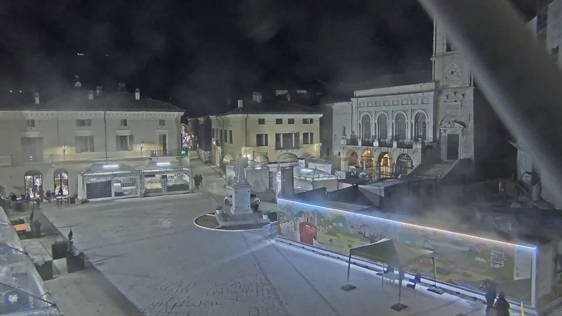 A nighttime view of a town square with several buildings, temporary market stalls, and a statue, under a hazy or foggy sky.