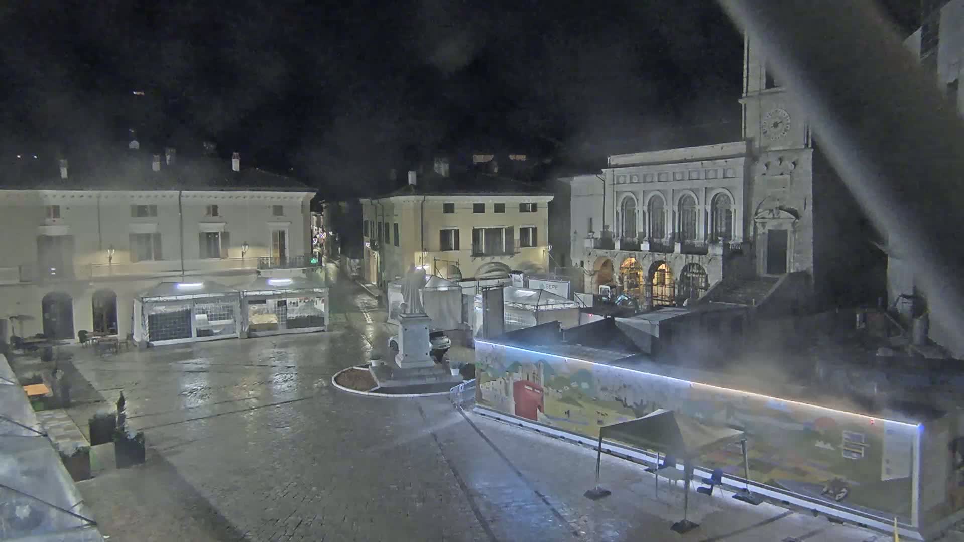 A wet, nighttime town square features several buildings, temporary market stalls, and a statue at its center.