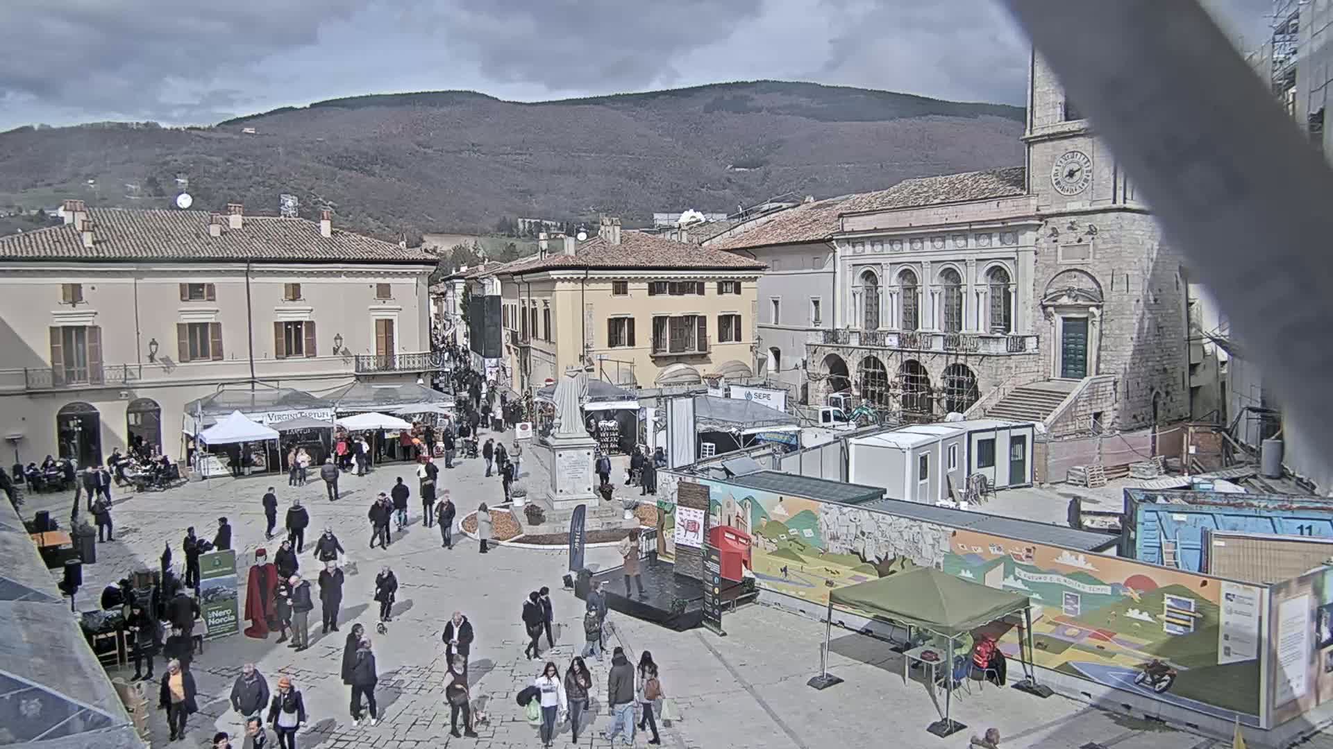 A town square filled with people and market stalls under a cloudy sky, with mountains in the background.