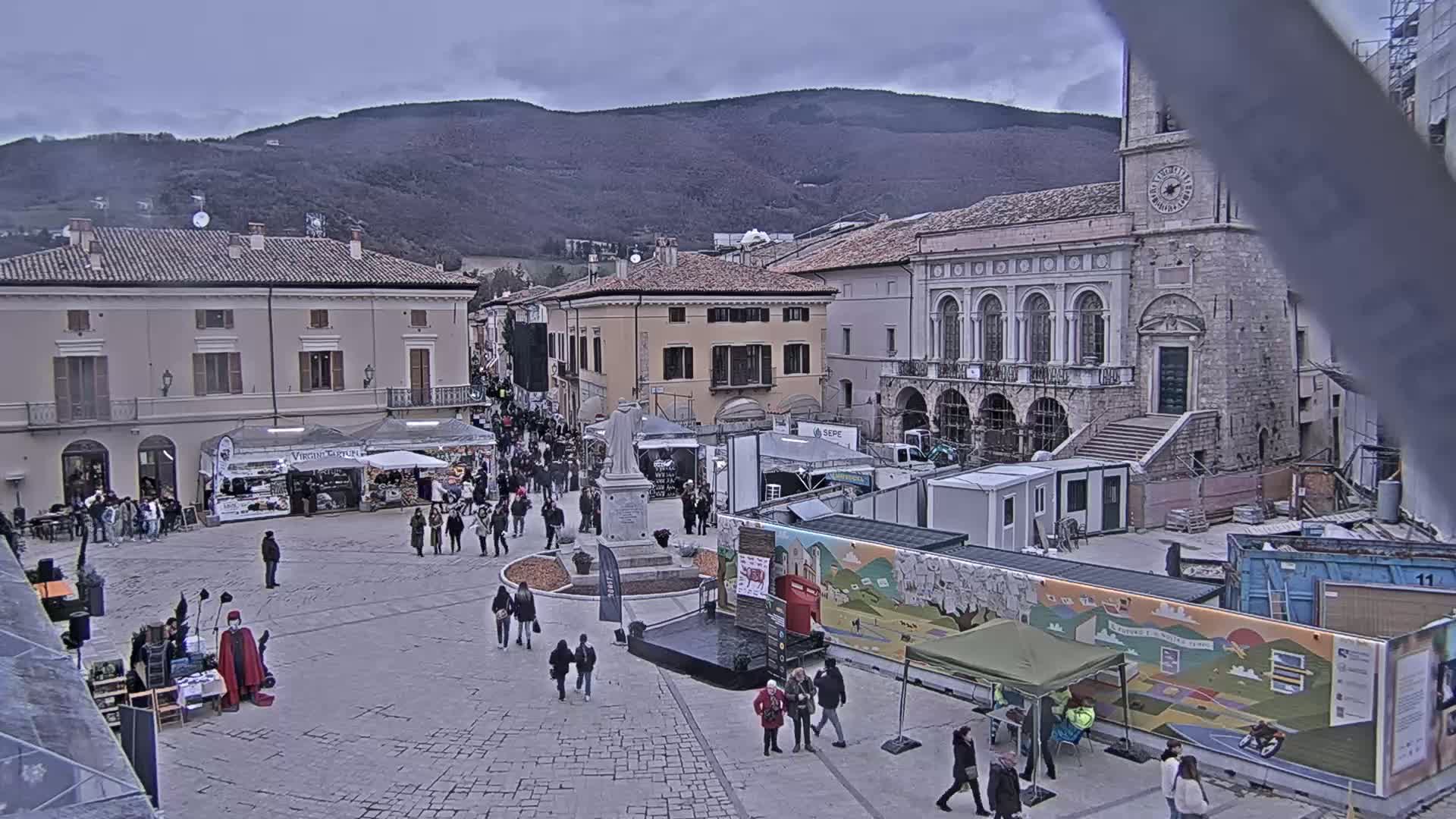 A town square with many people, market stalls, and buildings, under an overcast sky with mountains in the background.