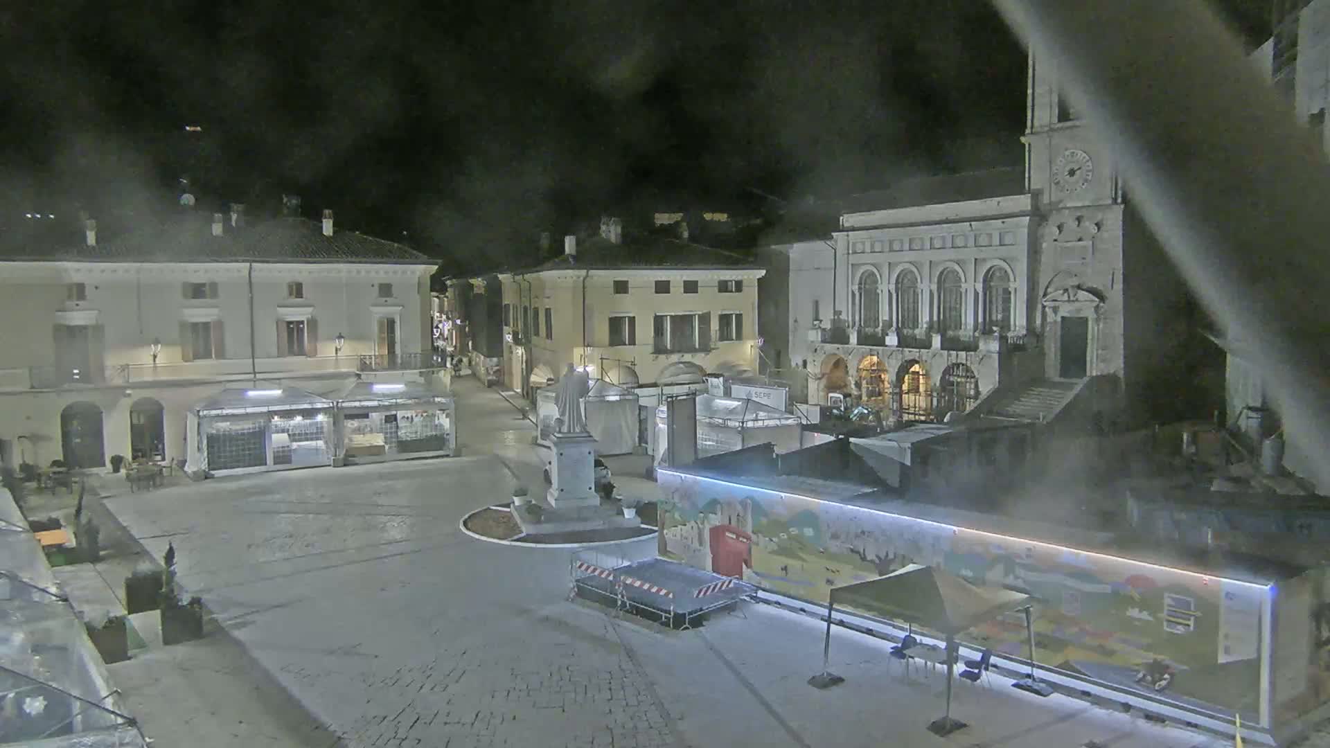 A nighttime view of a town square with several buildings, temporary market stalls, and a statue, under hazy or foggy conditions.