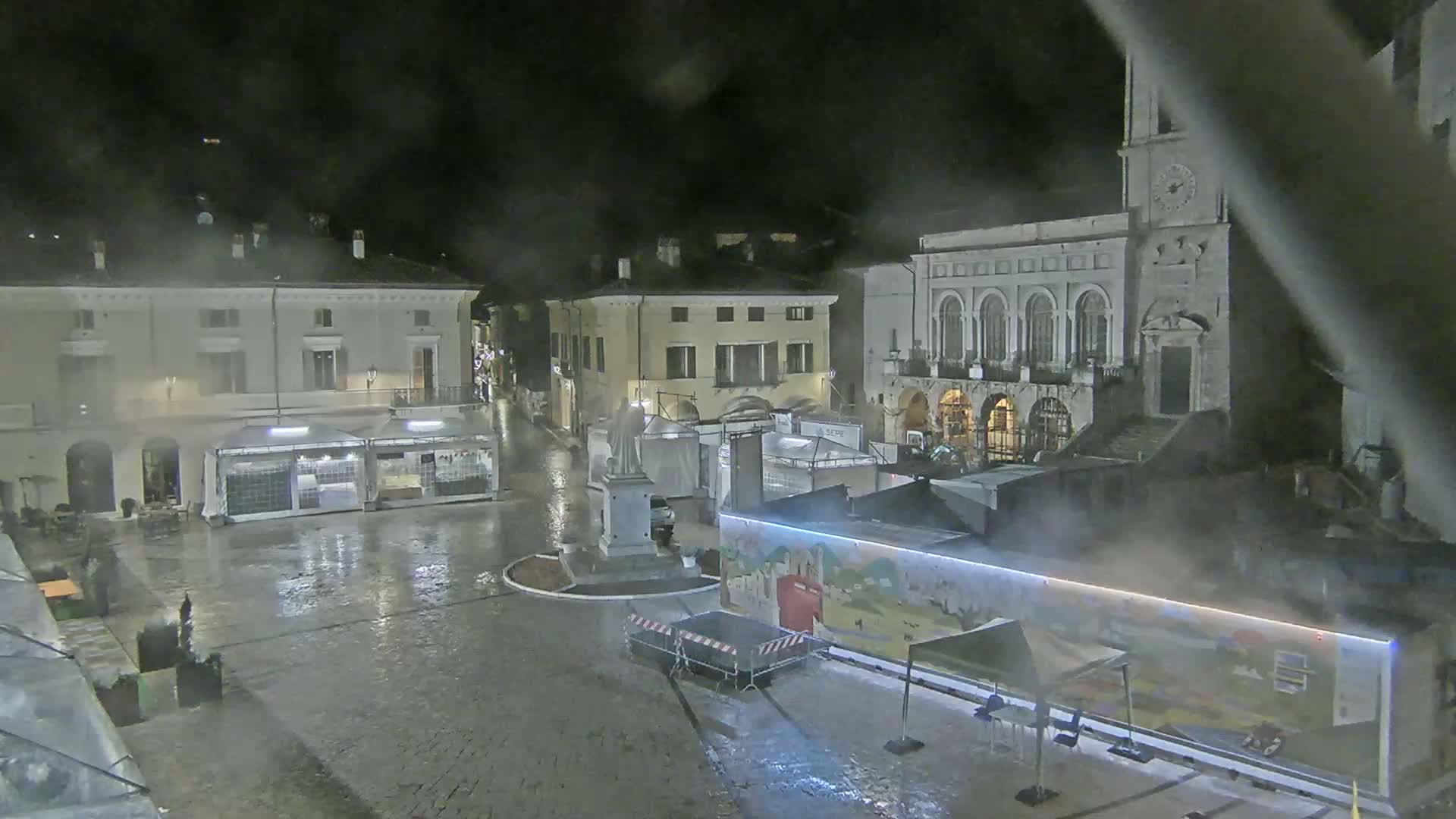 A rain-soaked town square at night, surrounded by buildings, with temporary structures and a statue in the center.