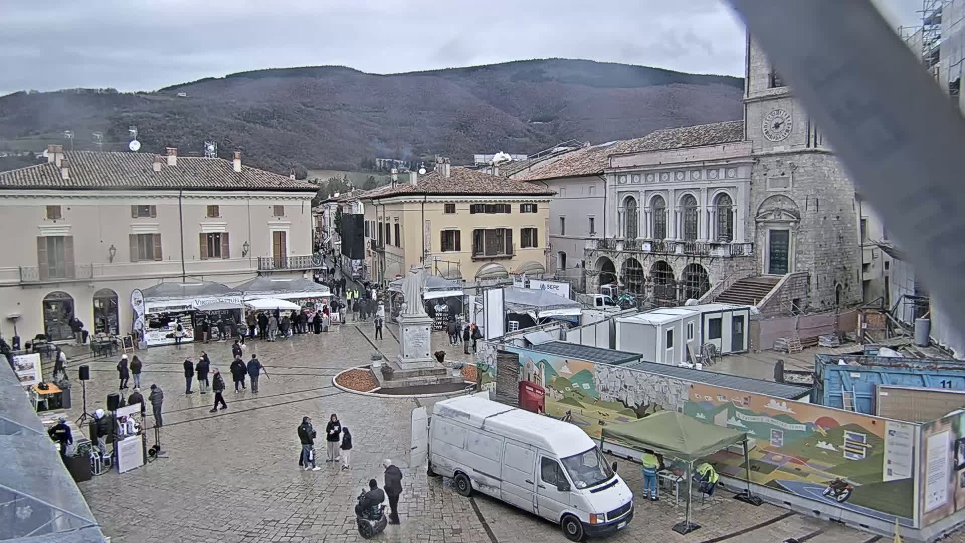 A town square with several temporary market stalls and vendors, a statue in the center, and various vehicles, under an overcast sky in front of older buildings and a mountain in the background.