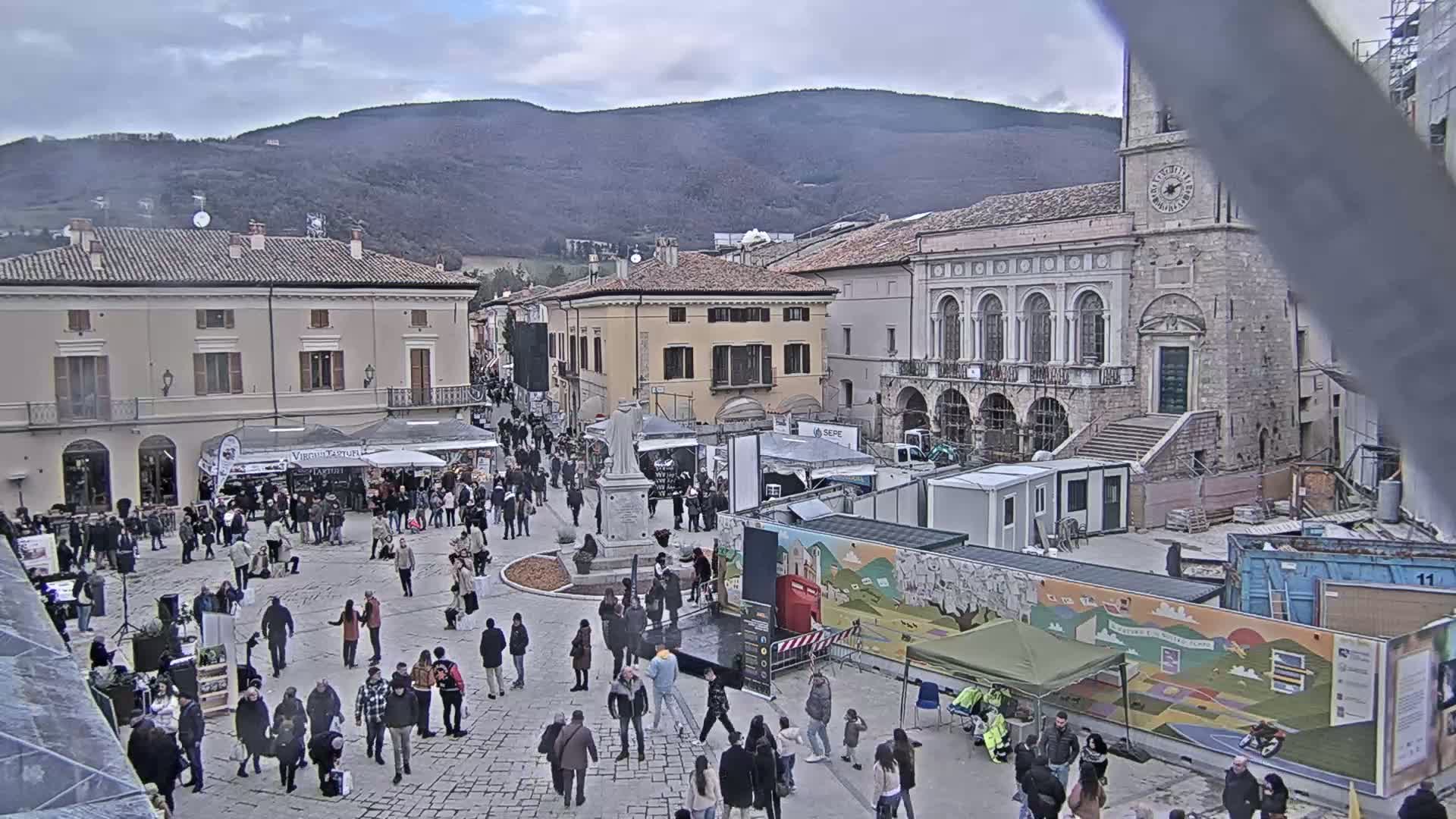 A town square filled with people and market stalls under an overcast sky, with mountains in the background and construction underway near a large building.
